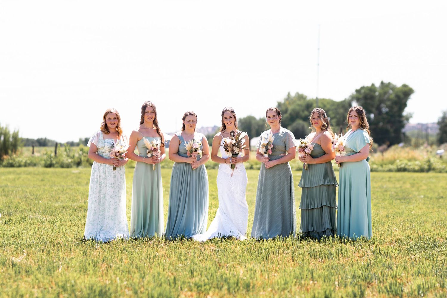 Bridal party in flowing pastel blue dresses pose together in a sunny meadow during an outdoor wedding ceremony.