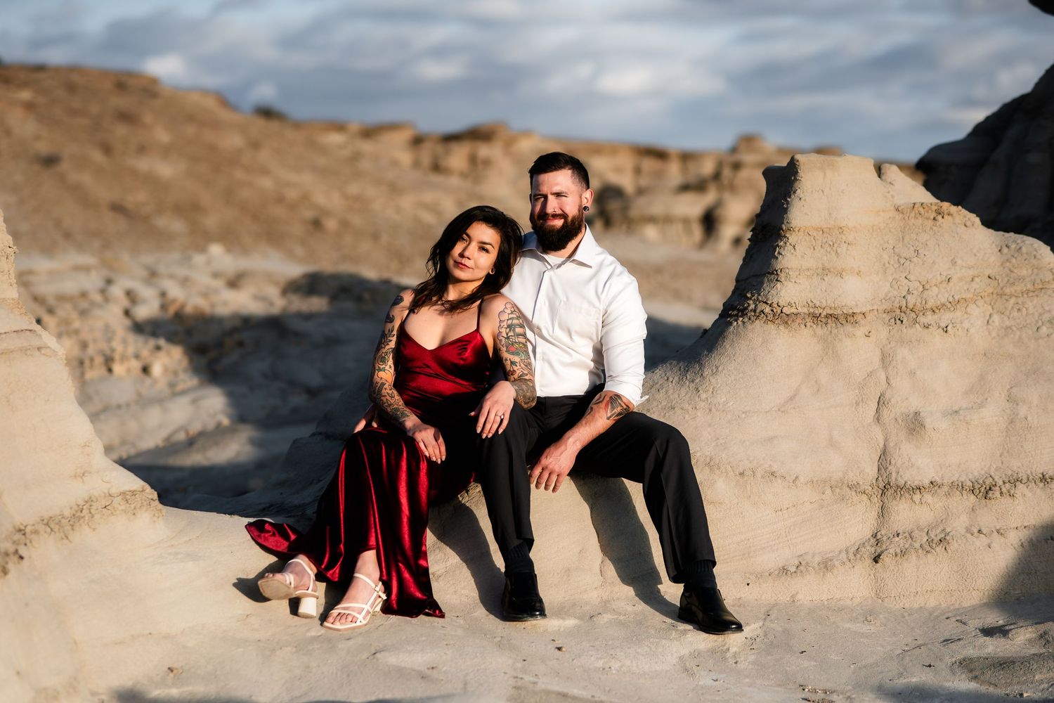 A couple poses together on desert rock formations with the woman wearing a flowing red dress.