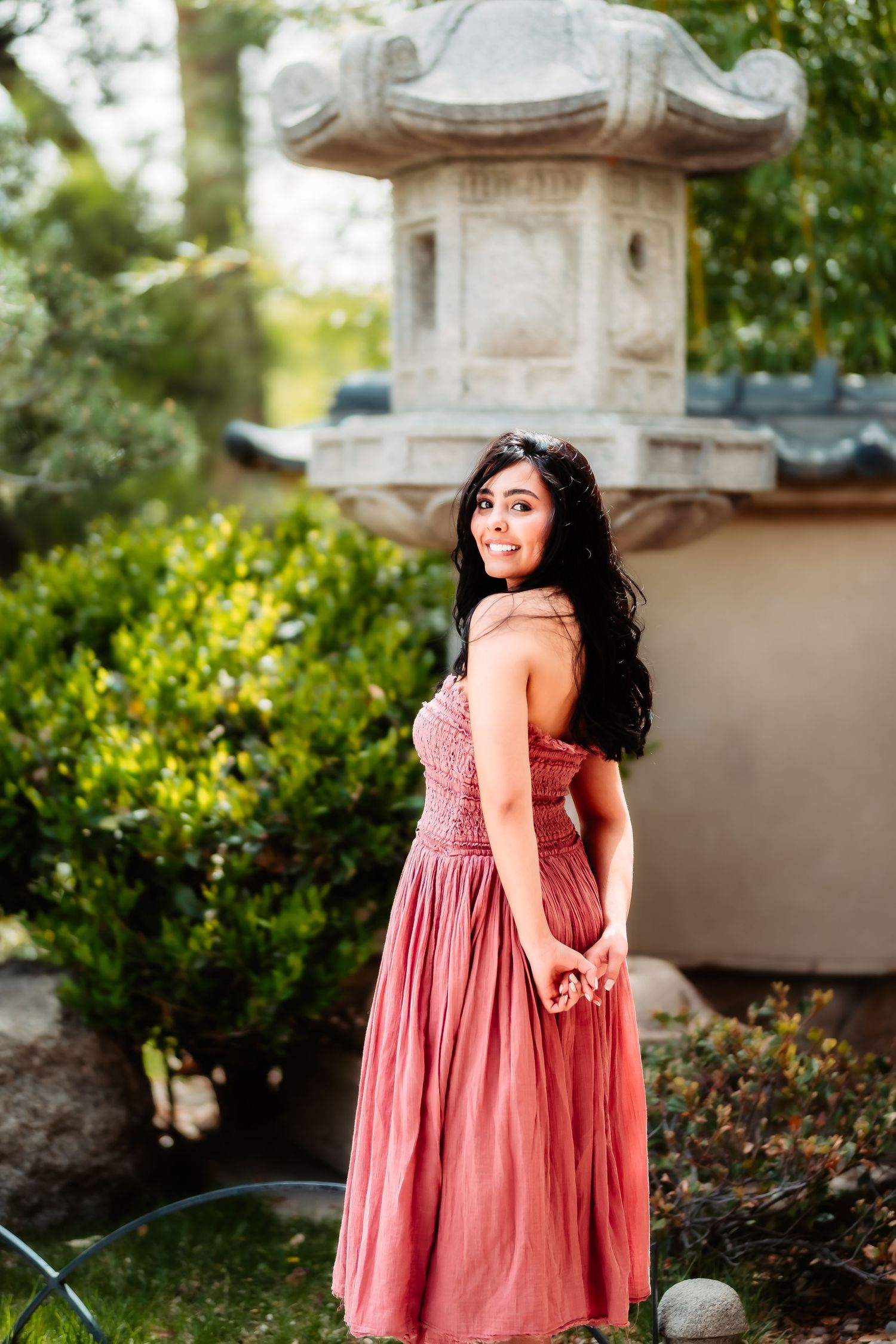 A woman in a pink maxi dress poses in front of an ornate stone garden lantern.