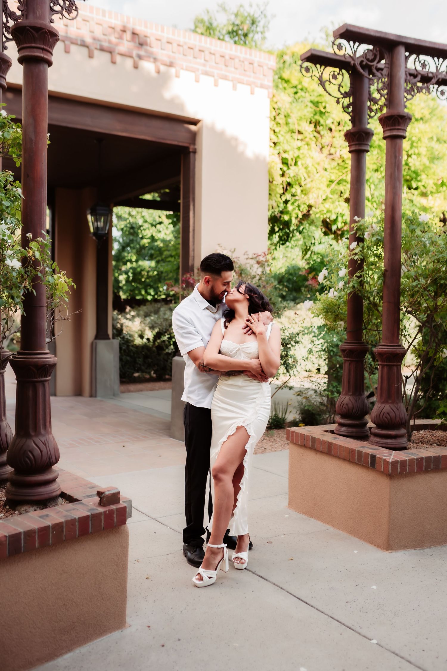 A couple dressed in white shares romantic moments under a covered walkway with decorative columns.