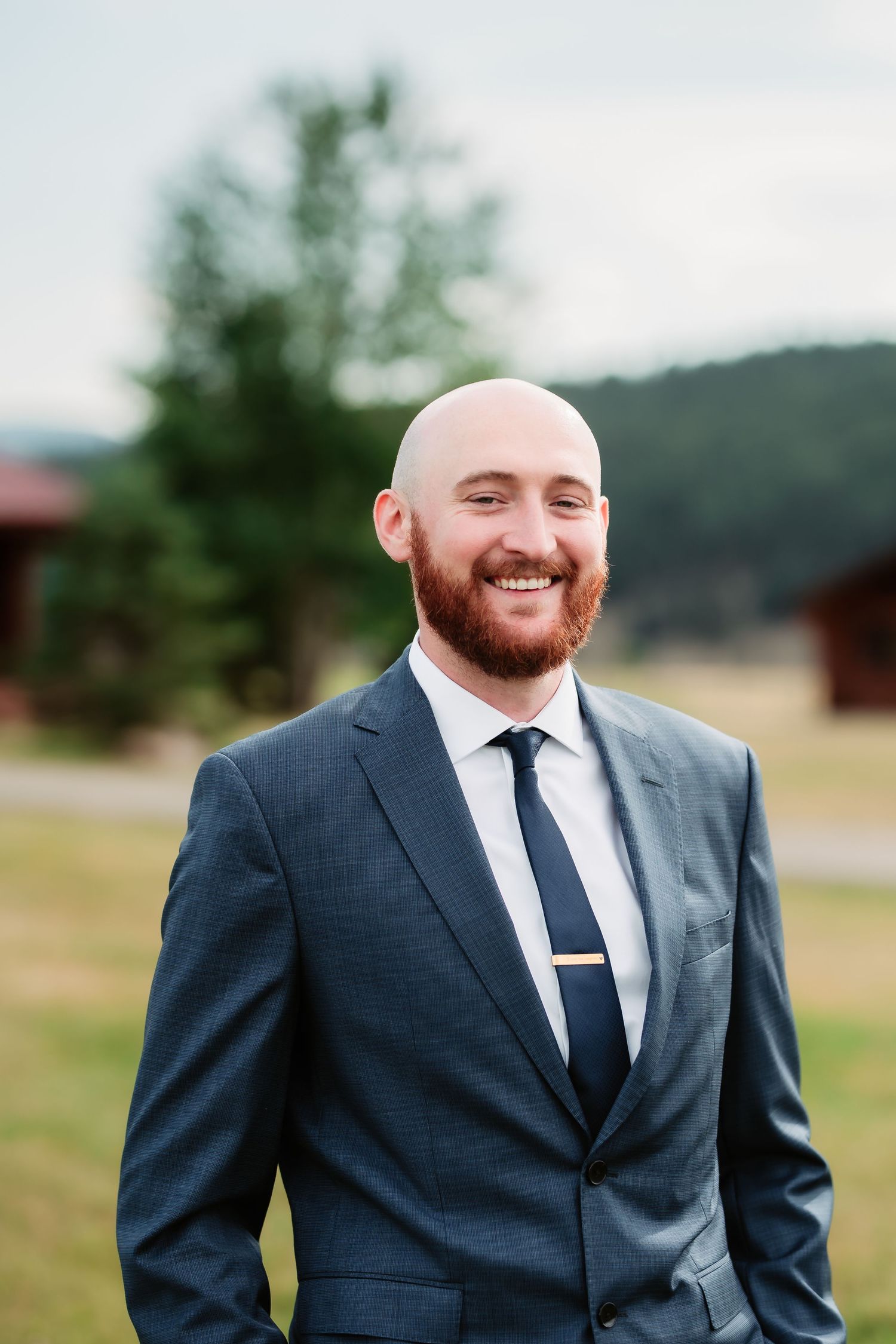 The groom wearing a navy suit and tie poses outdoors with mountains and trees in the background.