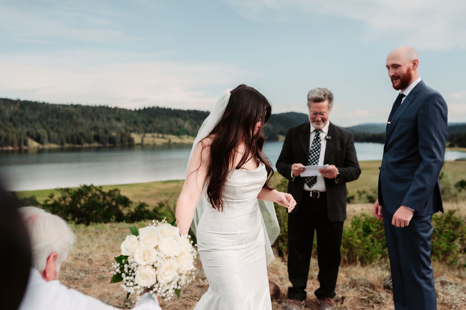An outdoor wedding ceremony unfolds against a scenic lake and mountain backdrop.