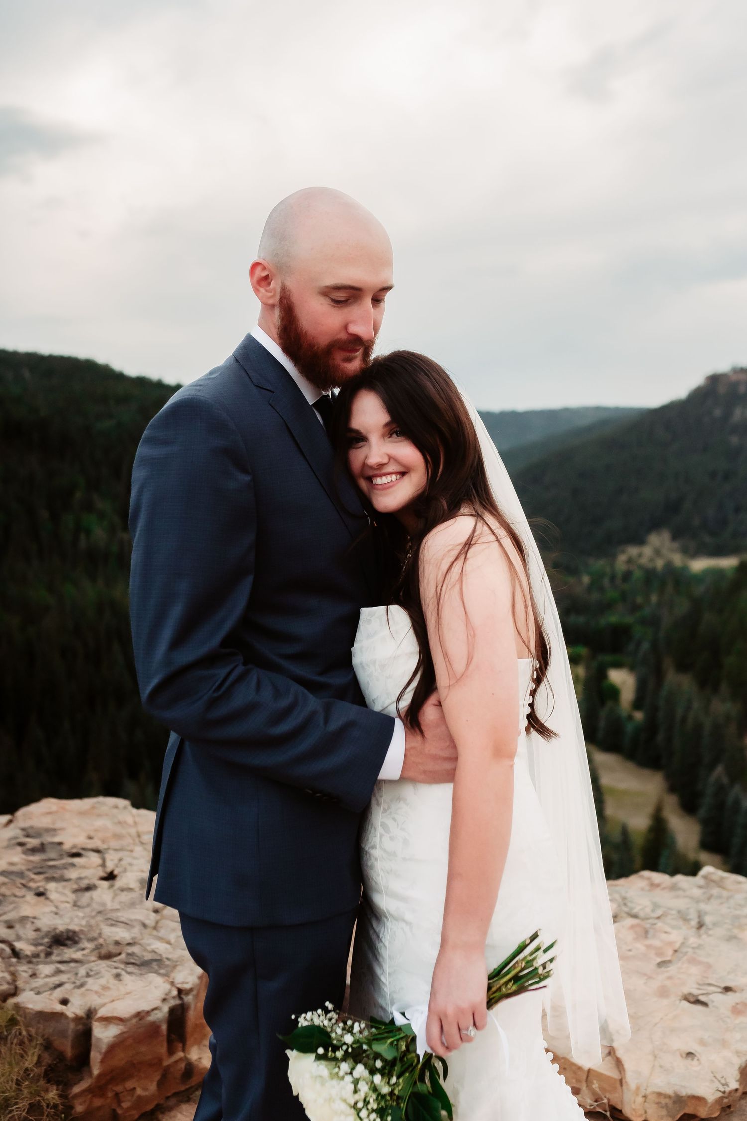 Romantic wedding photo of a couple embracing on a rocky clifftop with mountain views in the background at sunset.