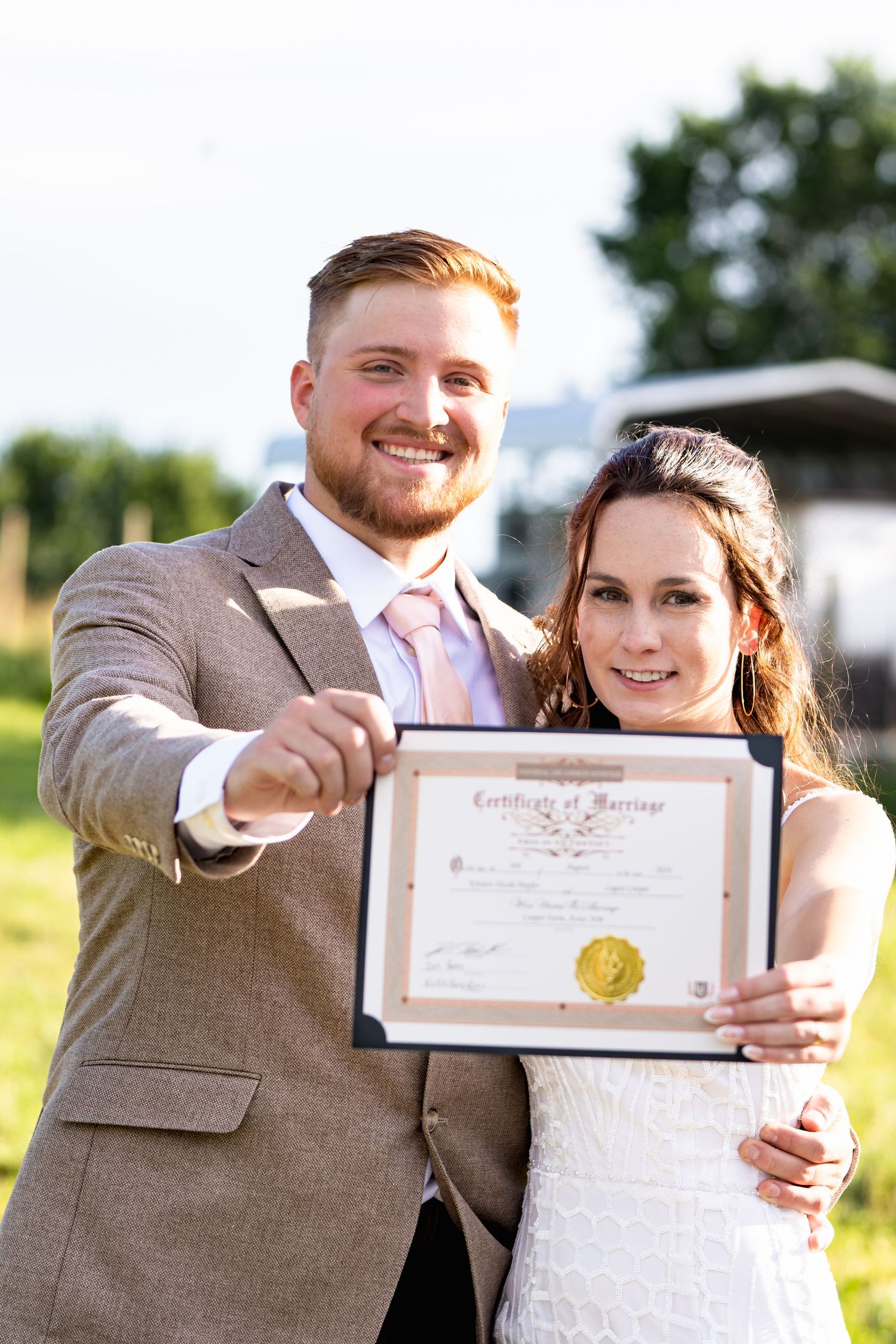 A smiling couple outdoors proudly holds up their marriage certificate with a golden seal.