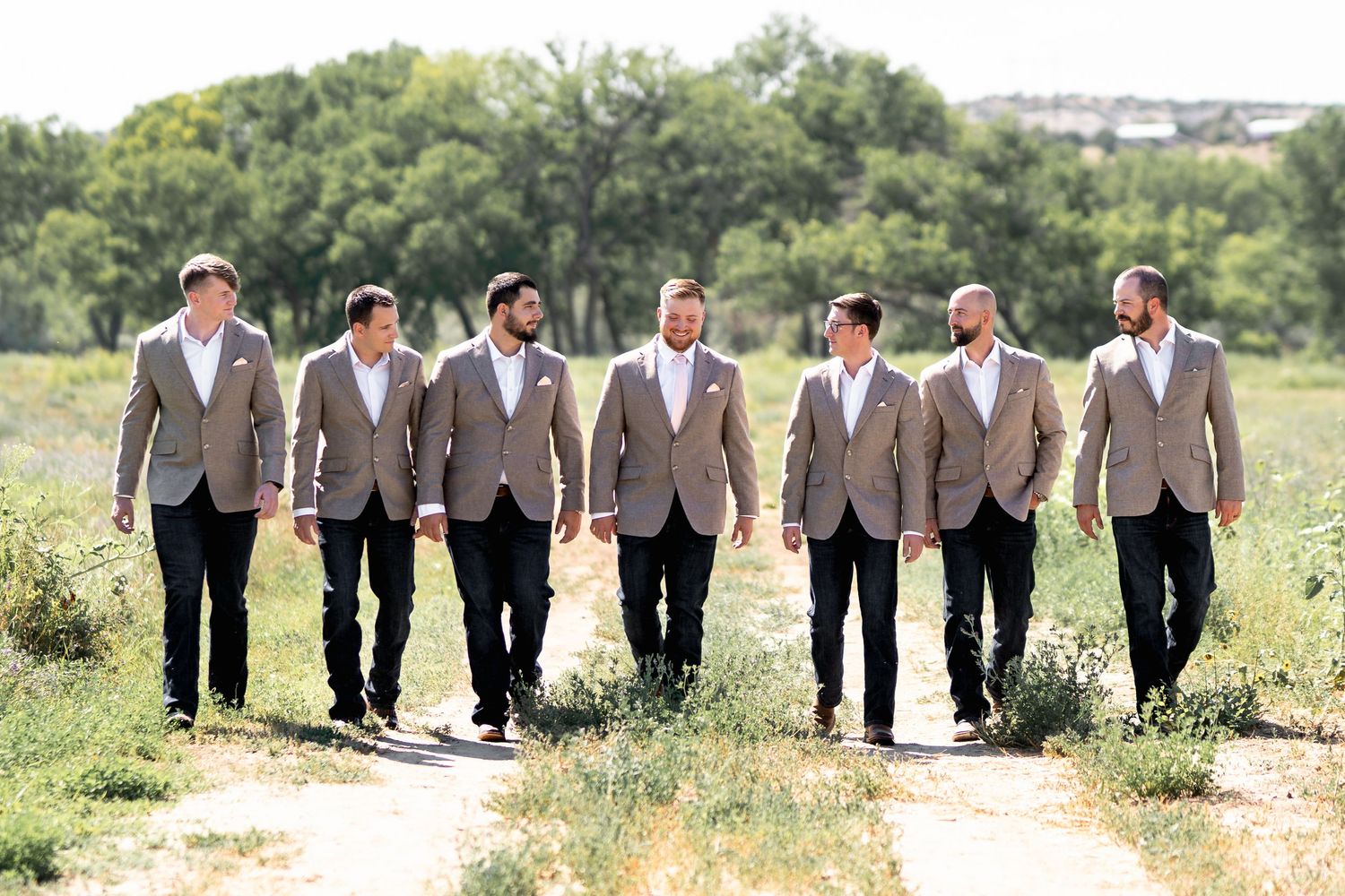 A group of seven groomsmen in matching tan blazers and dark pants walk together on a path through a grassy field.