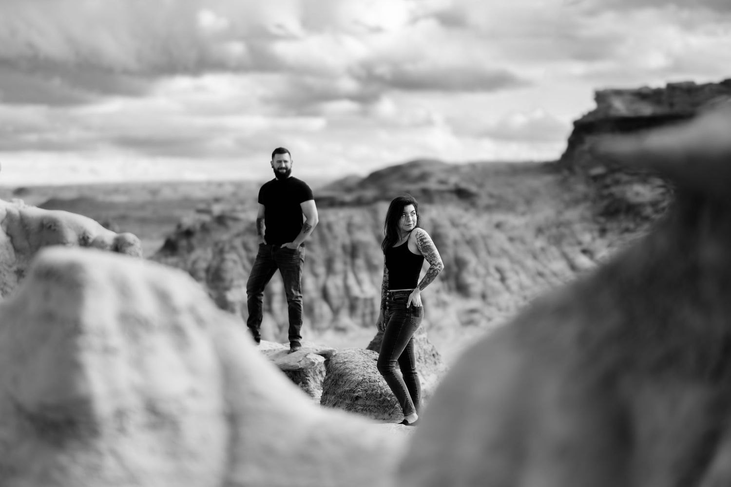 A black and white portrait of two people standing on dramatic rock formations in a desert landscape.