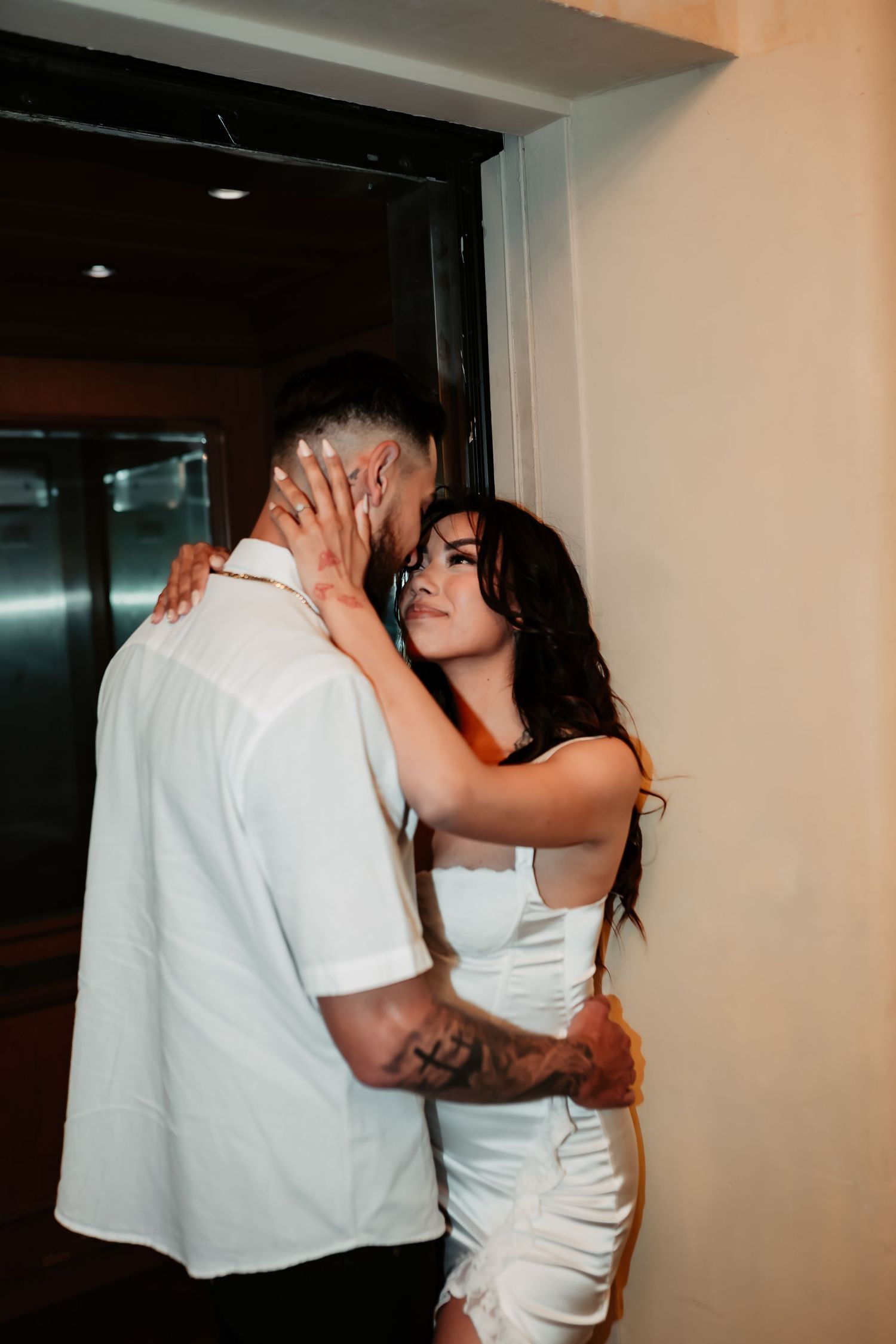 Romantic couple in white attire share an intimate moment near an elevator door.