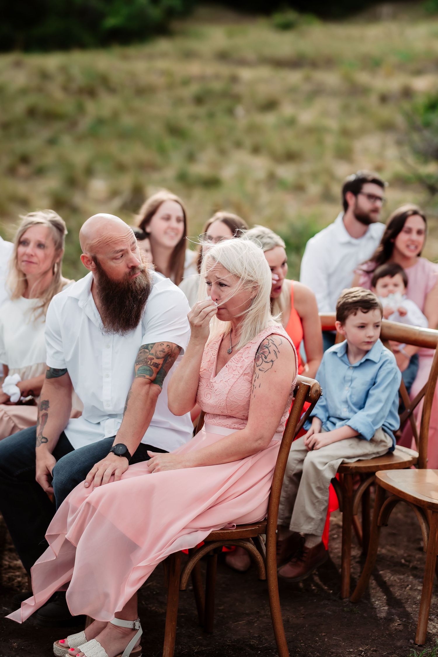Wedding guests seated in wooden chairs watch an outdoor ceremony against a mountainous backdrop.