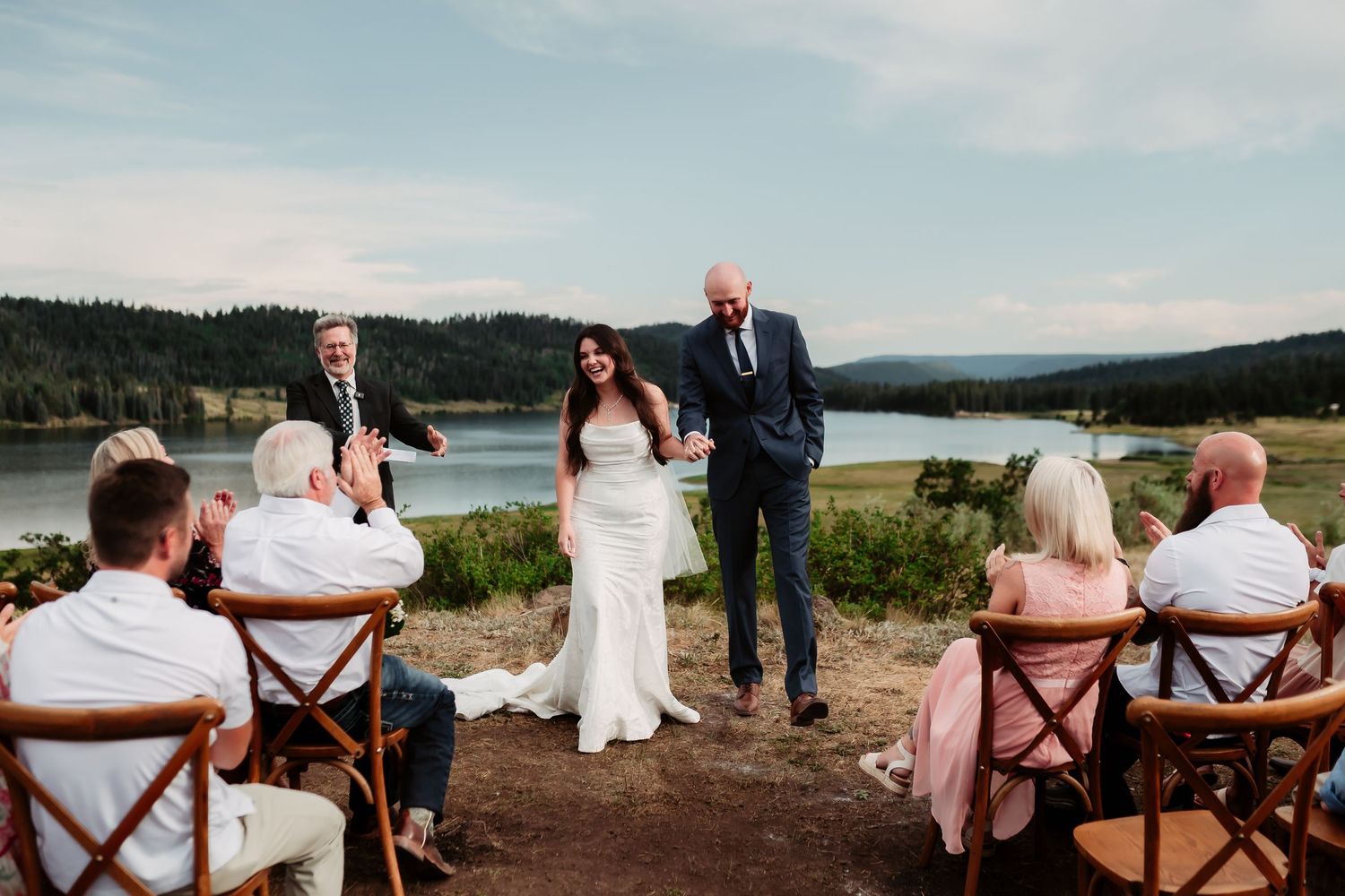 A lakeside wedding ceremony with guests seated in wooden chairs at sunset.