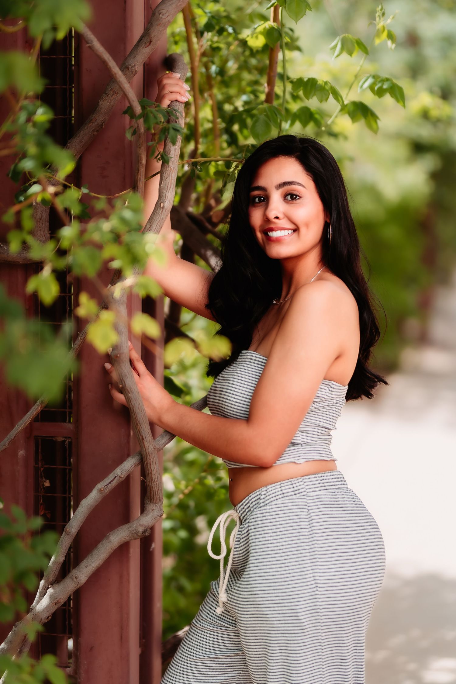 A woman in a striped two-piece outfit poses playfully near greenery and a wooden structure.