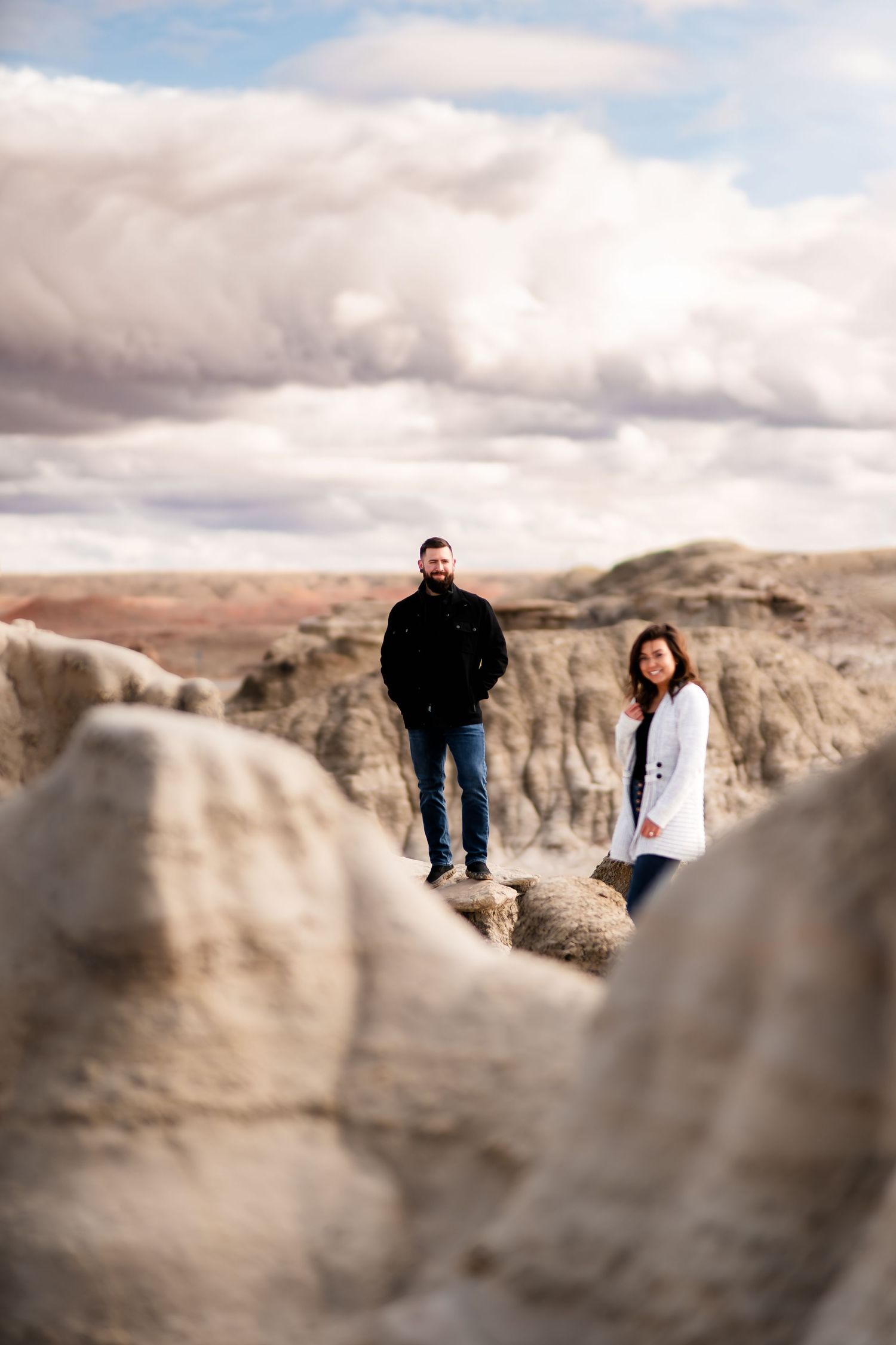 A couple stands on rocky terrain against a dramatic cloudy sky in a desert badlands landscape.