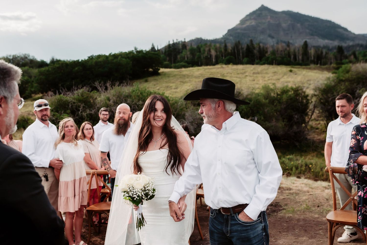 A wedding ceremony unfolds against a mountainous backdrop with guests seated on rustic chairs.