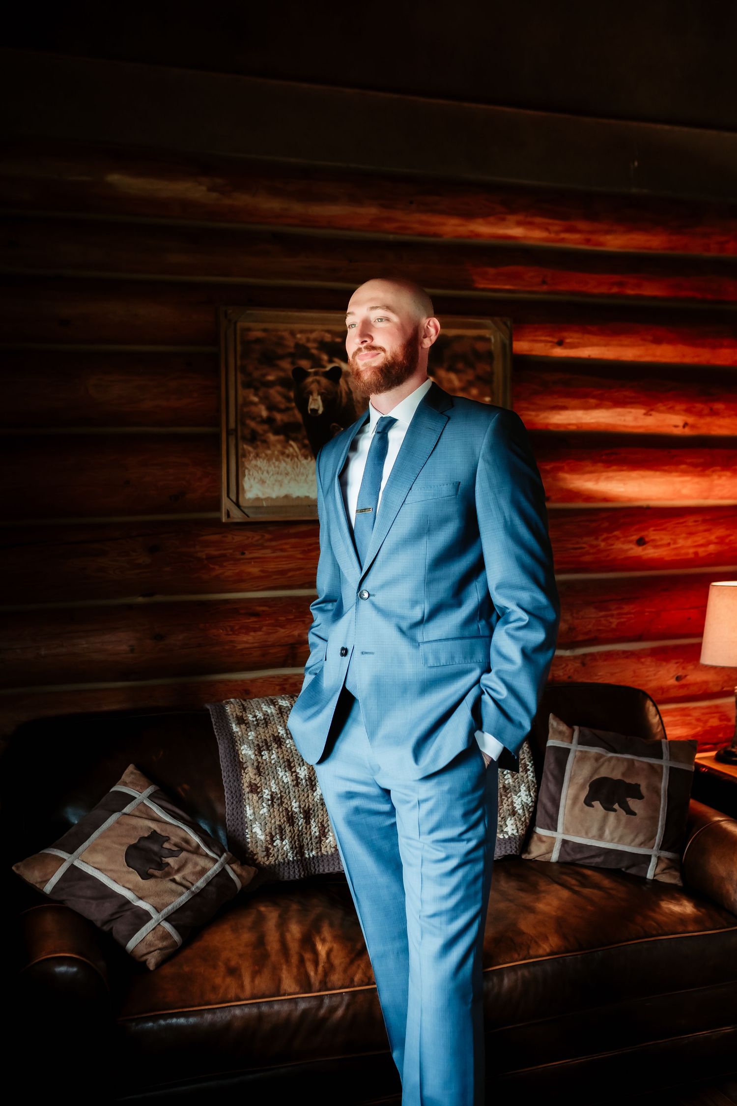 Groom wearing a powder blue suit poses against rustic wooden wall with decorative artwork.