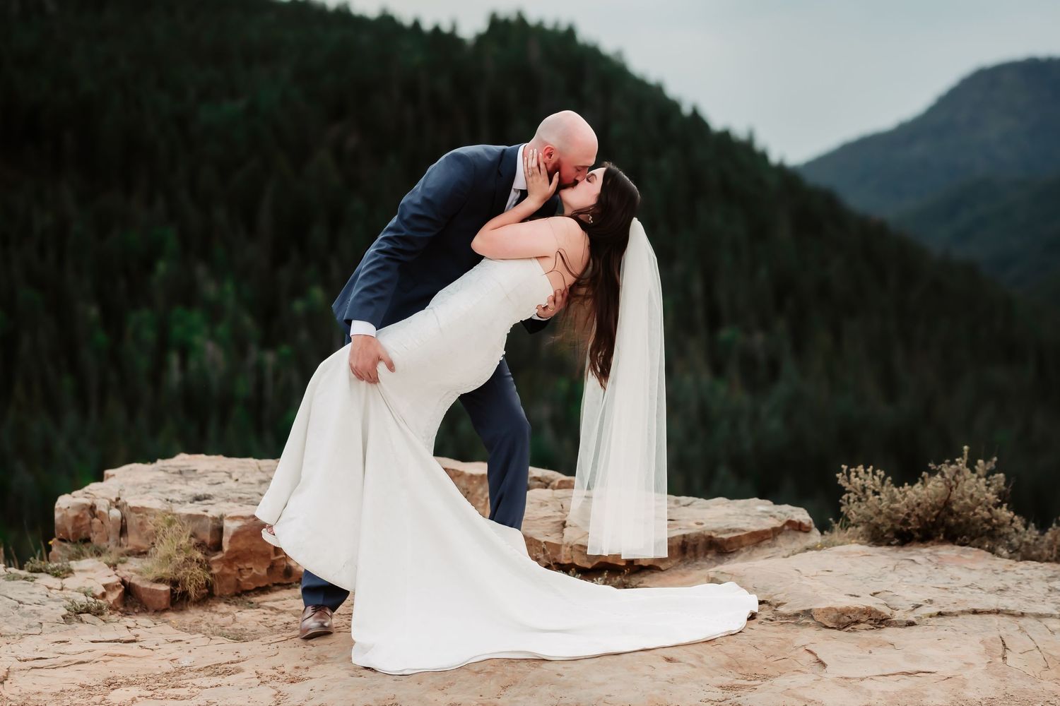 A romantic dip pose between newlyweds on a rocky mountain overlook.