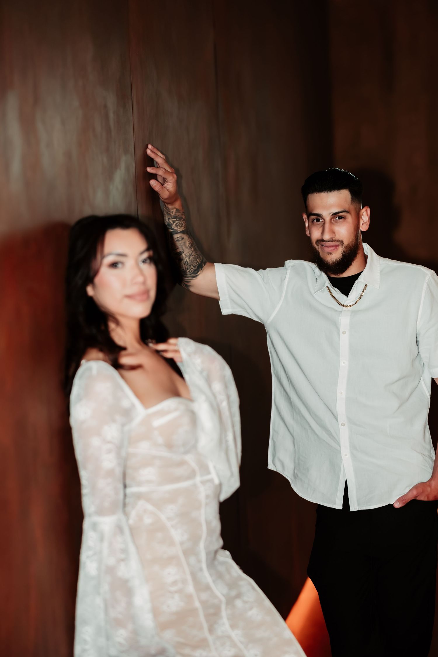 Couple in white outfits posing playfully against dark wall with dramatic lighting.
