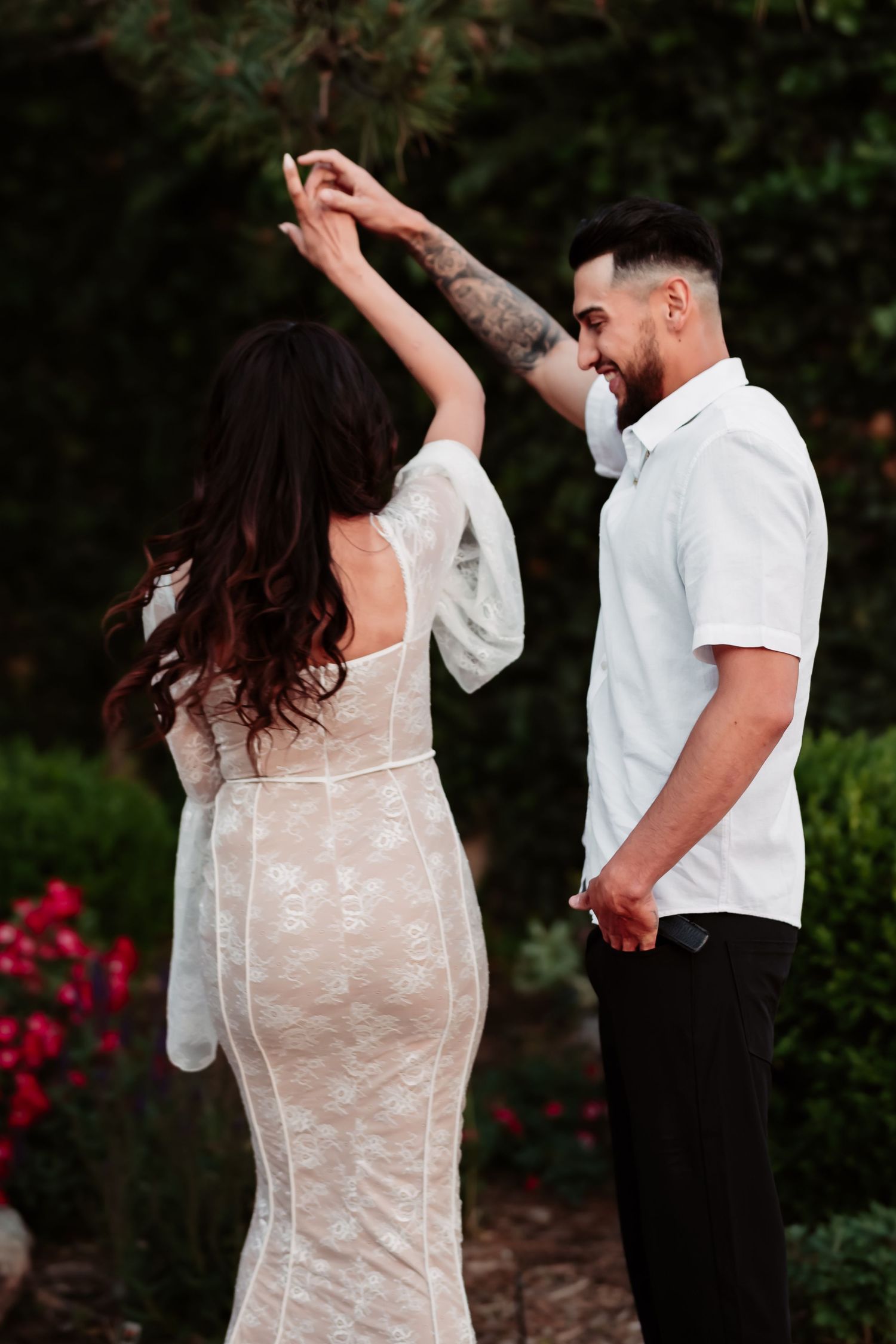 Couple in formal wear share a romantic dance moment in a garden with blooming red flowers.