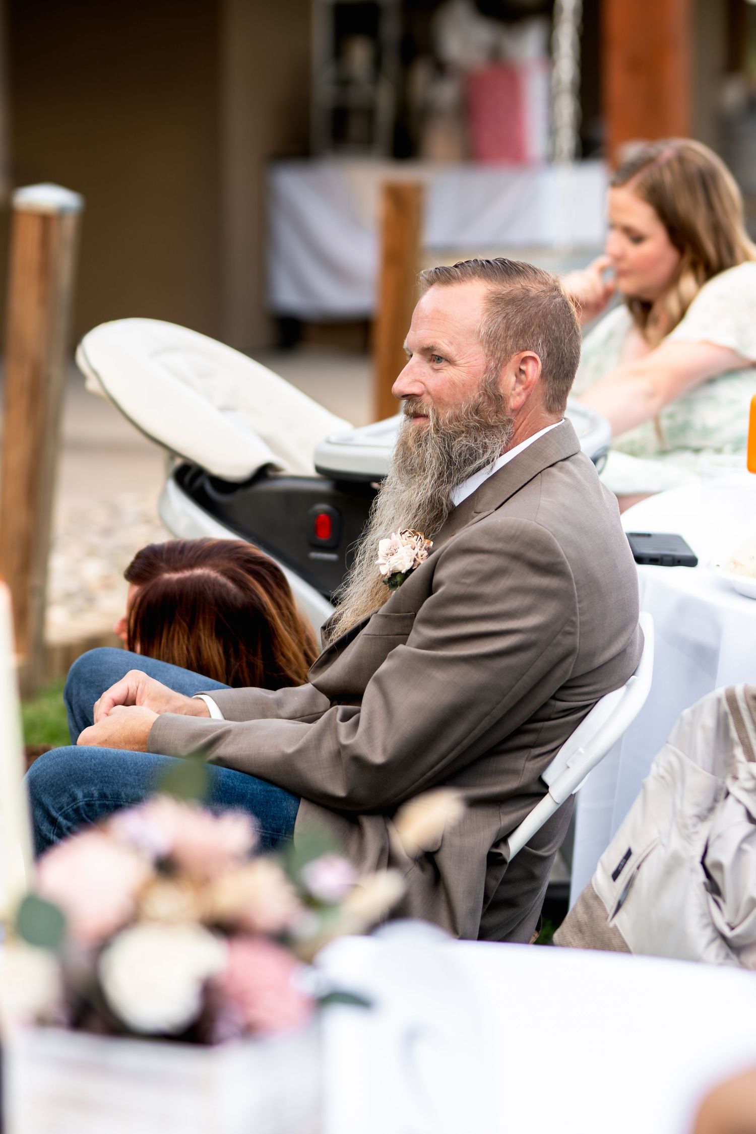 A guest in formal attire with a long beard watches the wedding ceremony from white folding chairs.
