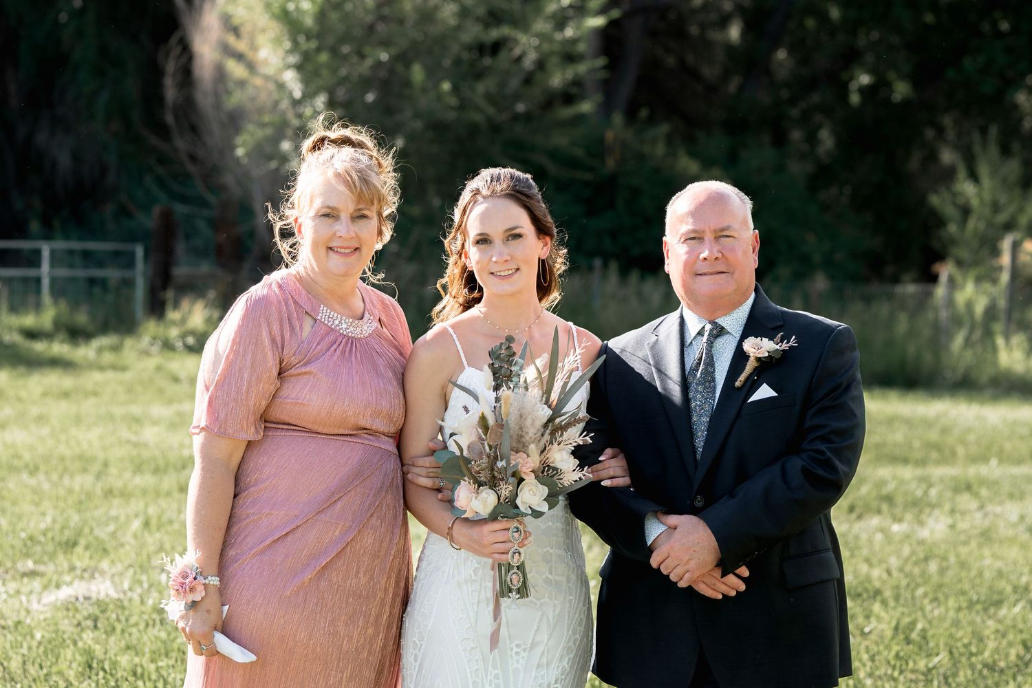 Three people in formal attire pose together outdoors at a wedding ceremony with a beautiful natural backdrop.