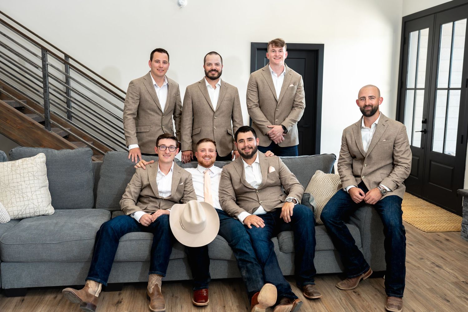 A seated group of groomsmen wearing coordinated tan suits gather for a formal wedding photo on a gray couch.