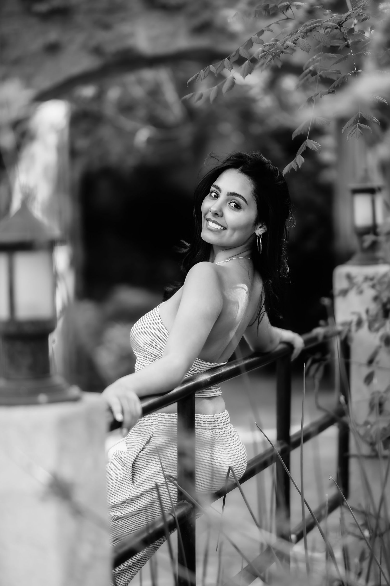 Black and white portrait showing a person in a striped dress posing by a railing with lanterns in the background.