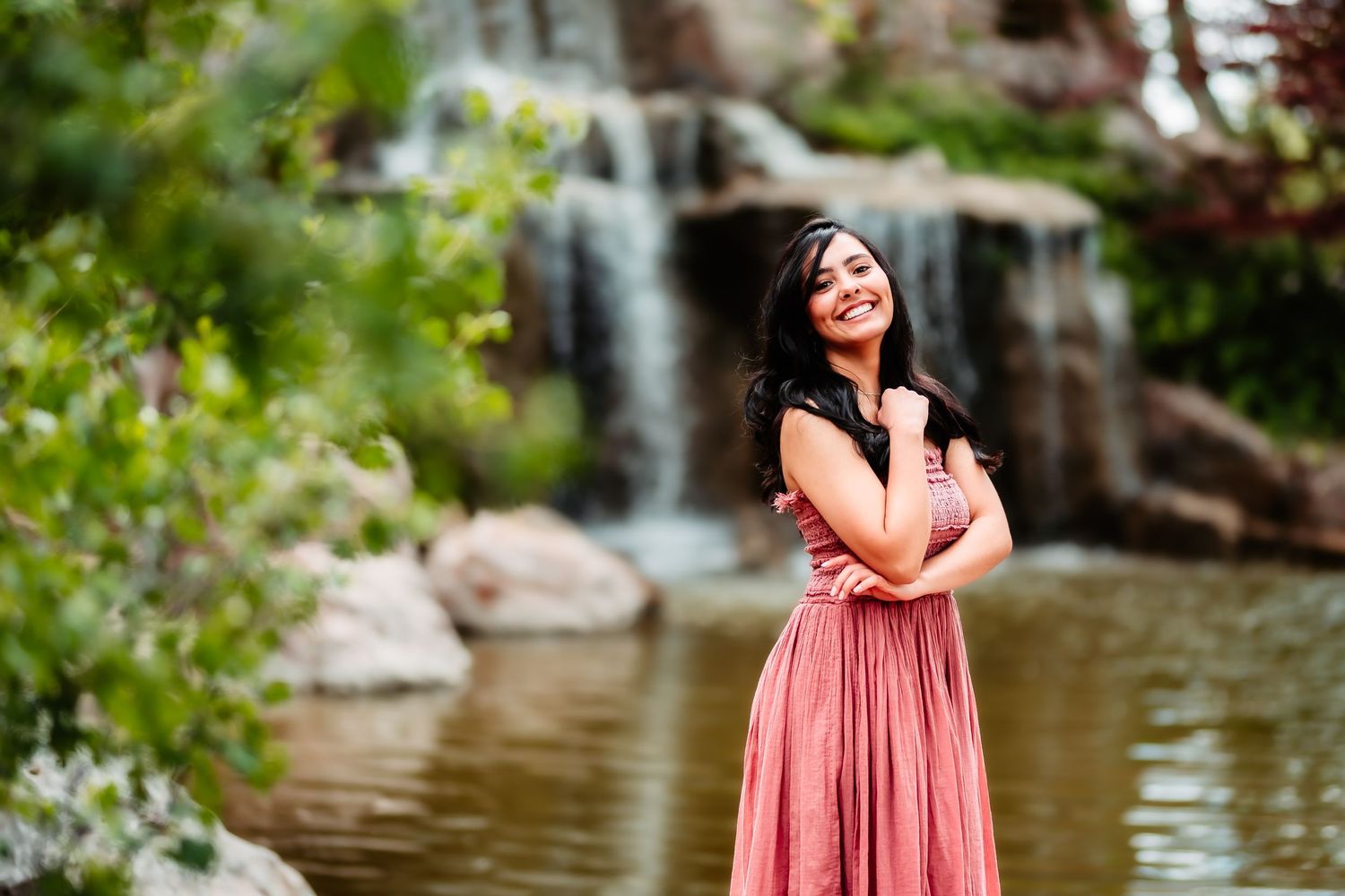 A person in a coral maxi dress poses by a scenic waterfall in a natural setting.