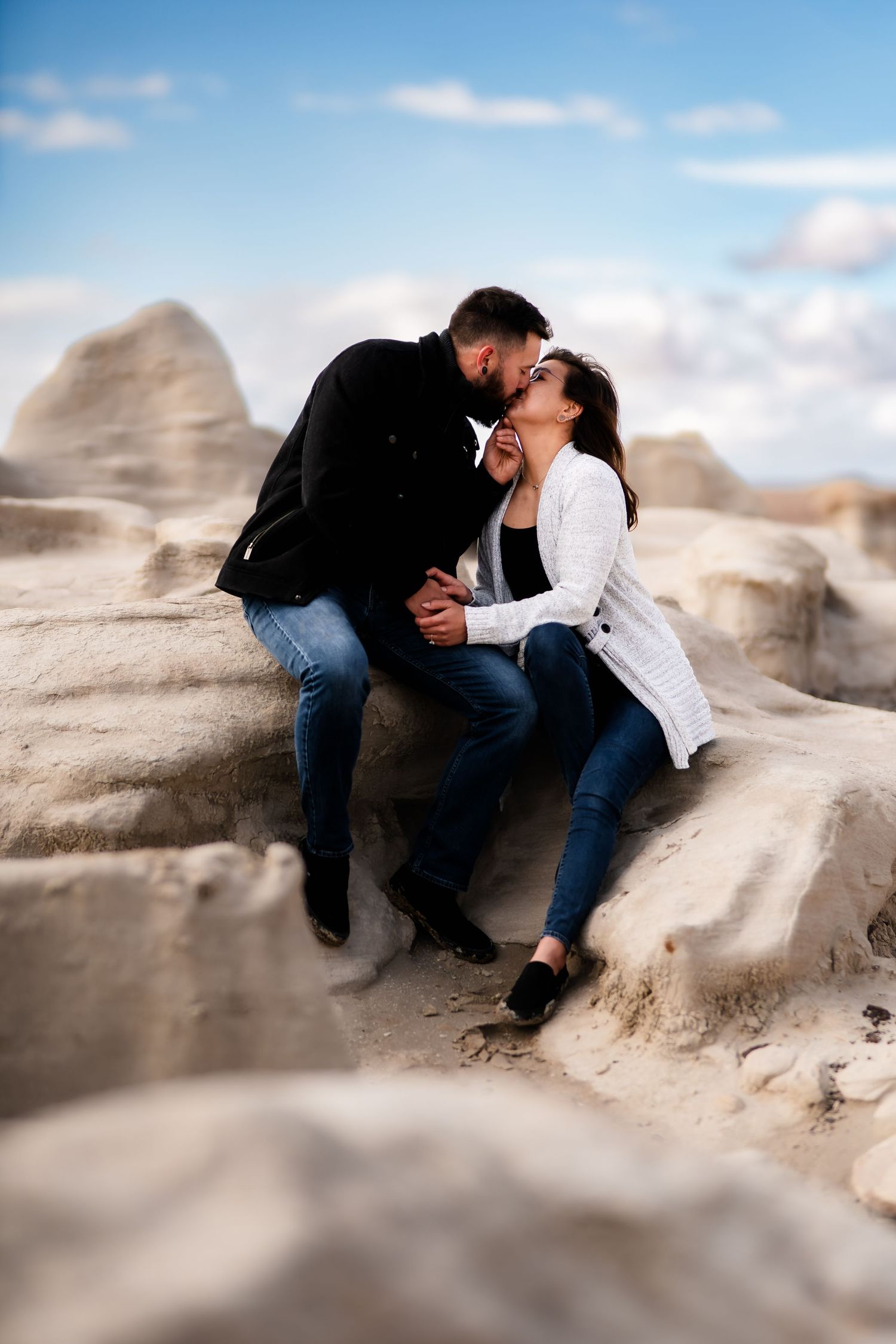 A romantic couple sharing an intimate moment while sitting on rocky desert terrain under a blue sky.