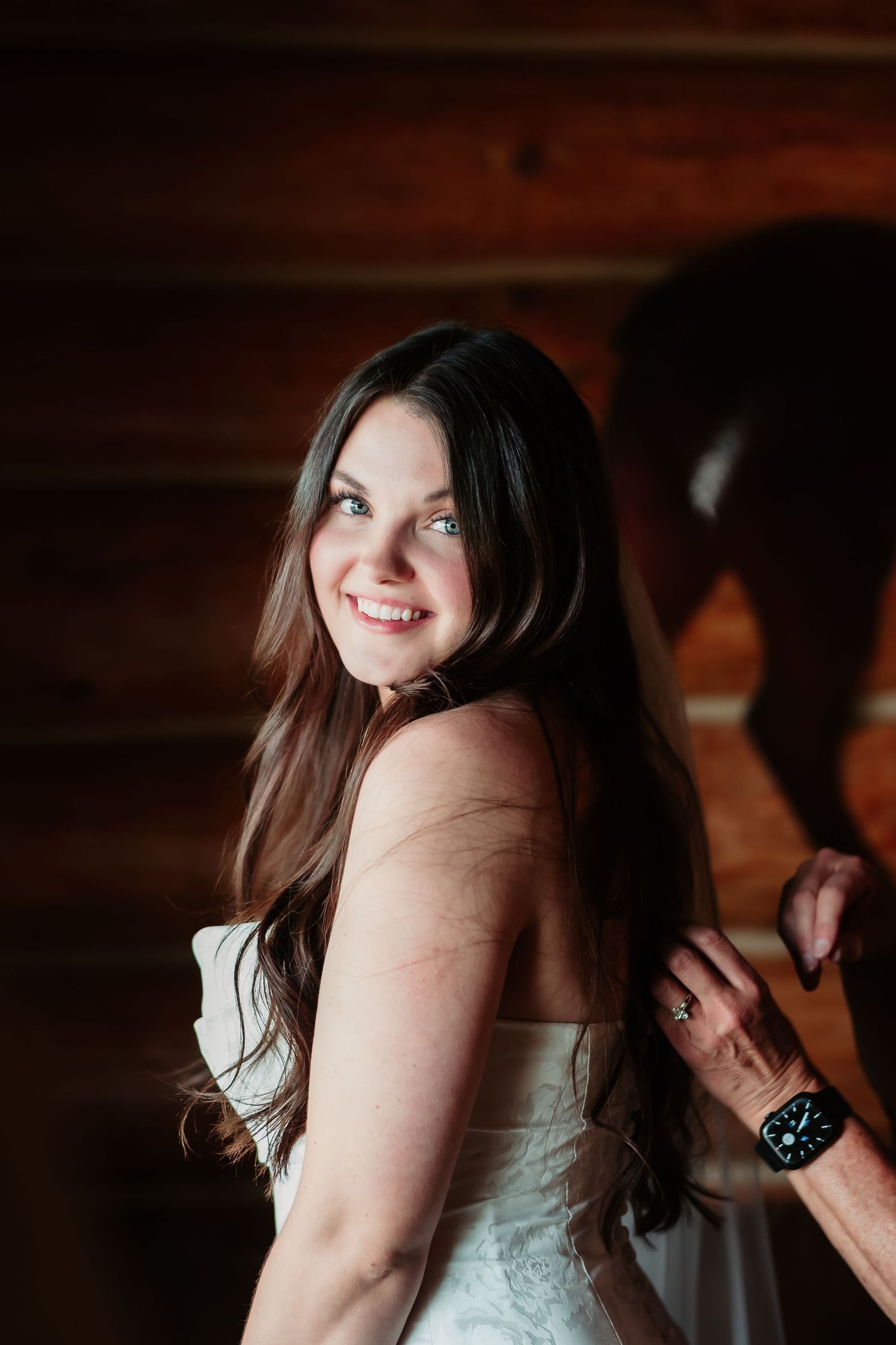 A bride in a white wedding gown smiles while getting ready in a rustic cabin setting.