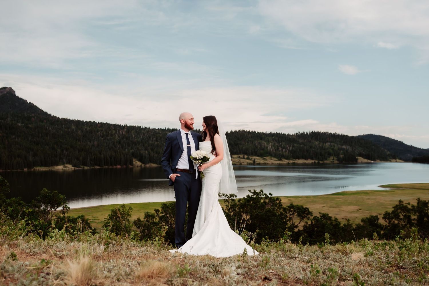 A couple shares a romantic moment overlooking a scenic lake and mountain landscape on their wedding day.
