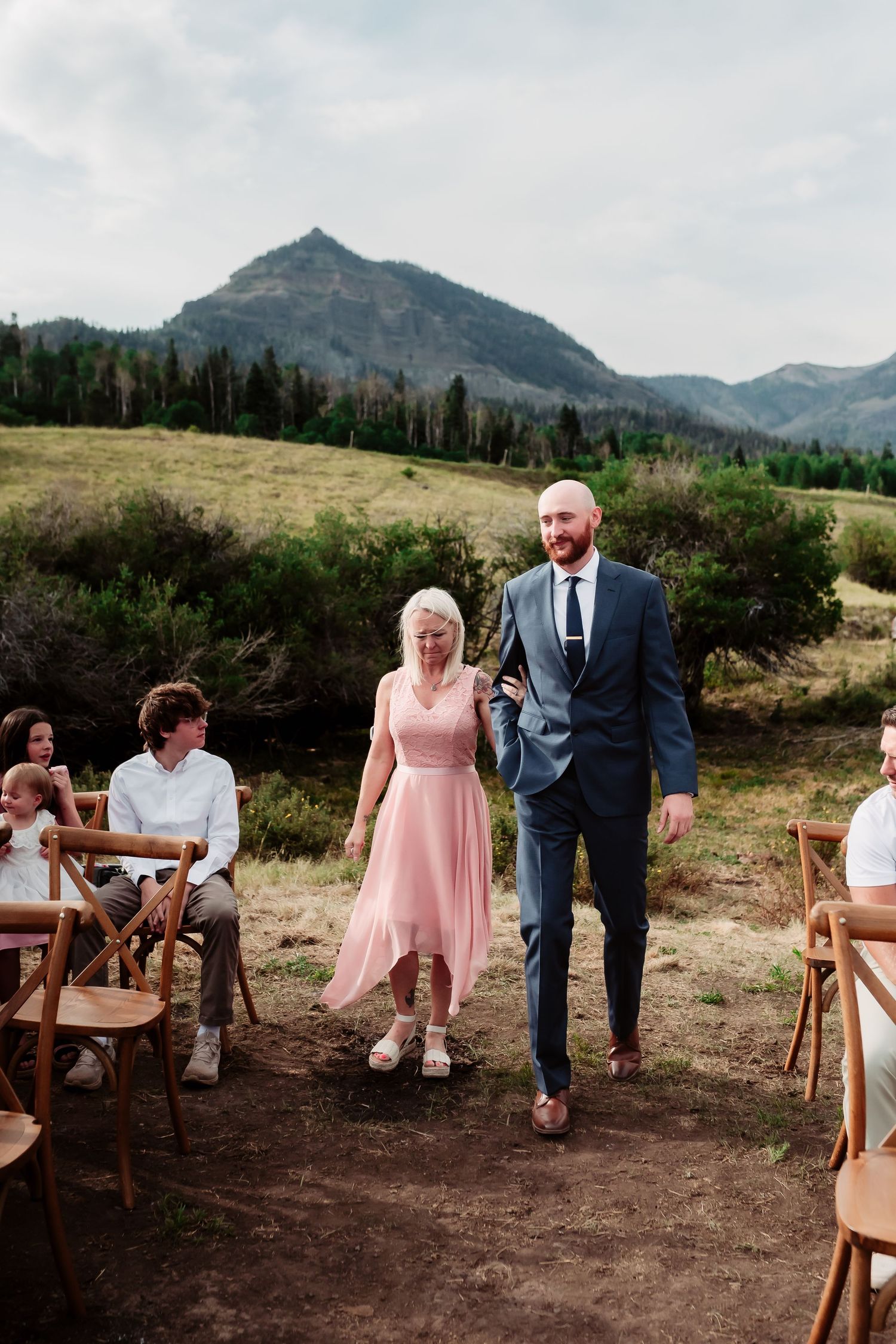 A couple walks down the aisle at an outdoor mountain wedding ceremony.