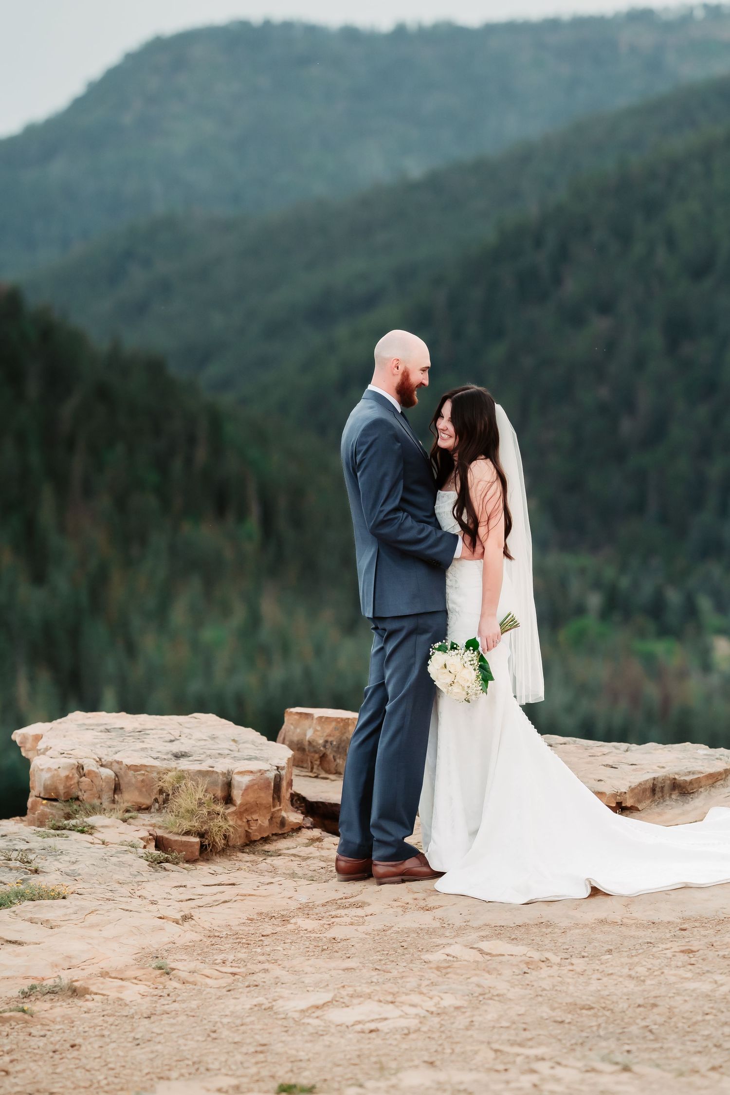 Wedding couple poses on rocky mountain overlook with dramatic forest valley backdrop.