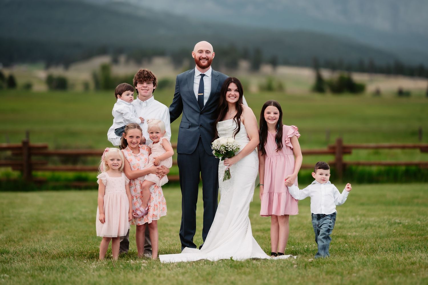 A wedding party poses together in a grassy field with mountains in the background.