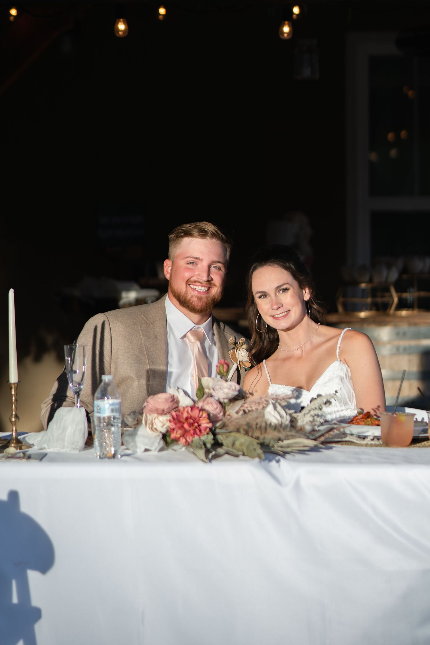 A couple sits at an elegantly decorated reception table with candlelight and floral centerpieces.