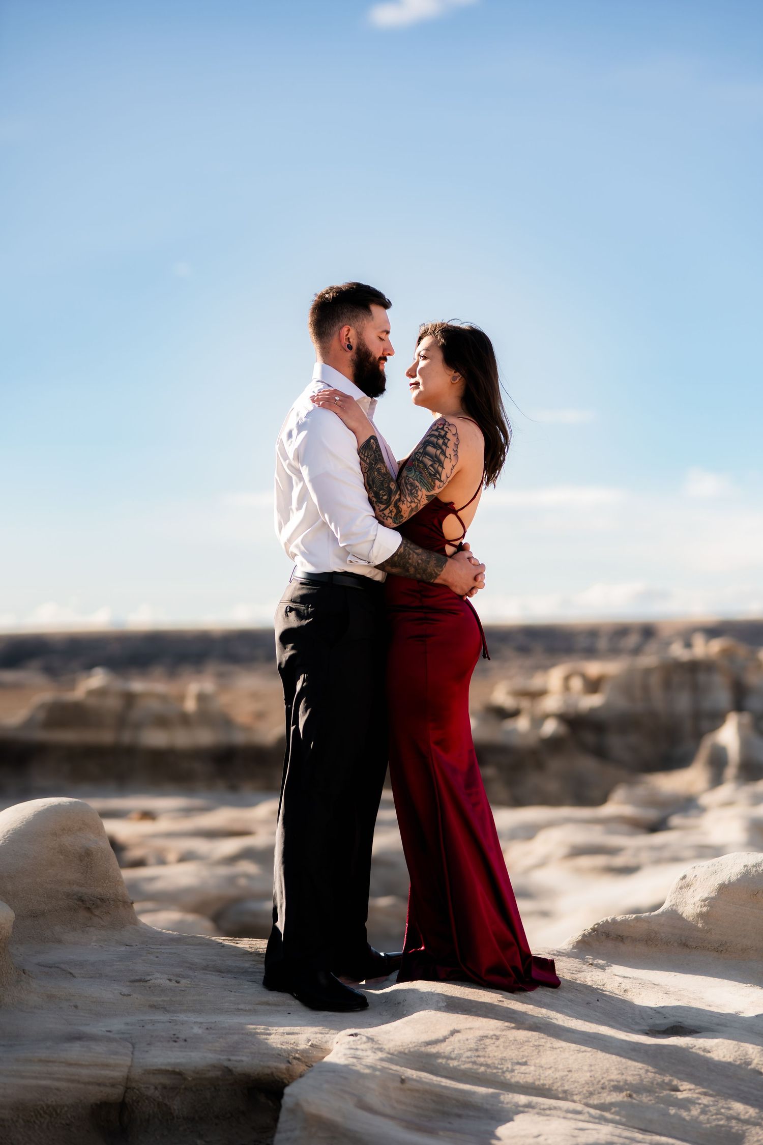 A couple shares an intimate moment on rocky terrain, with her wearing a red dress and him in formal attire.