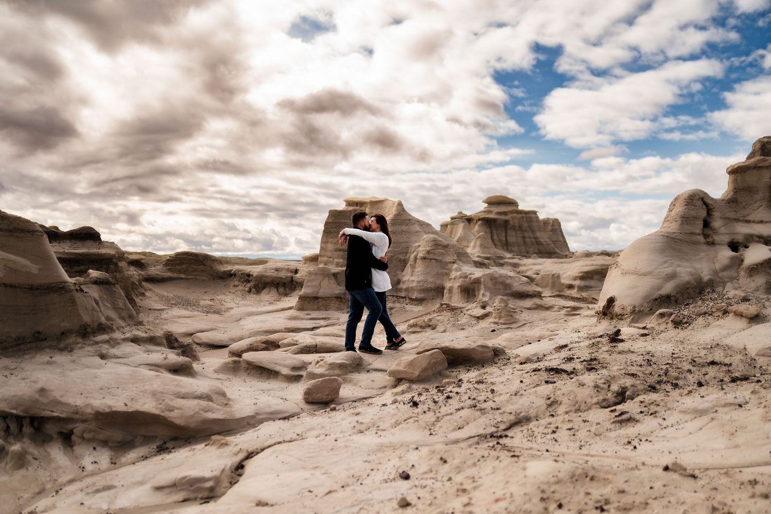 A silhouette of a couple stands on dramatic rock formations in a desert landscape under cloudy skies at sunset.