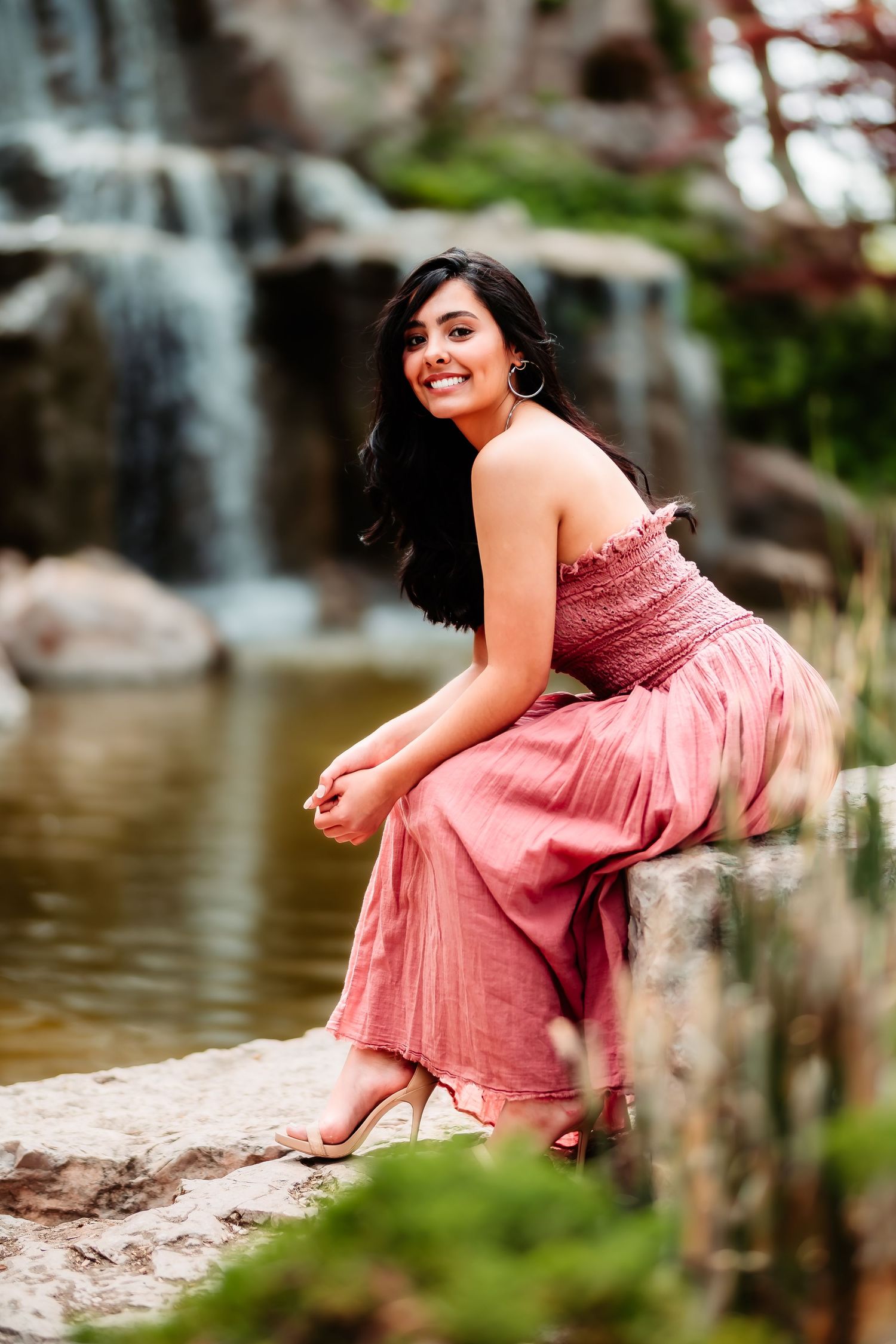 Woman in pink maxi dress sitting by waterfall on rocks in garden setting.