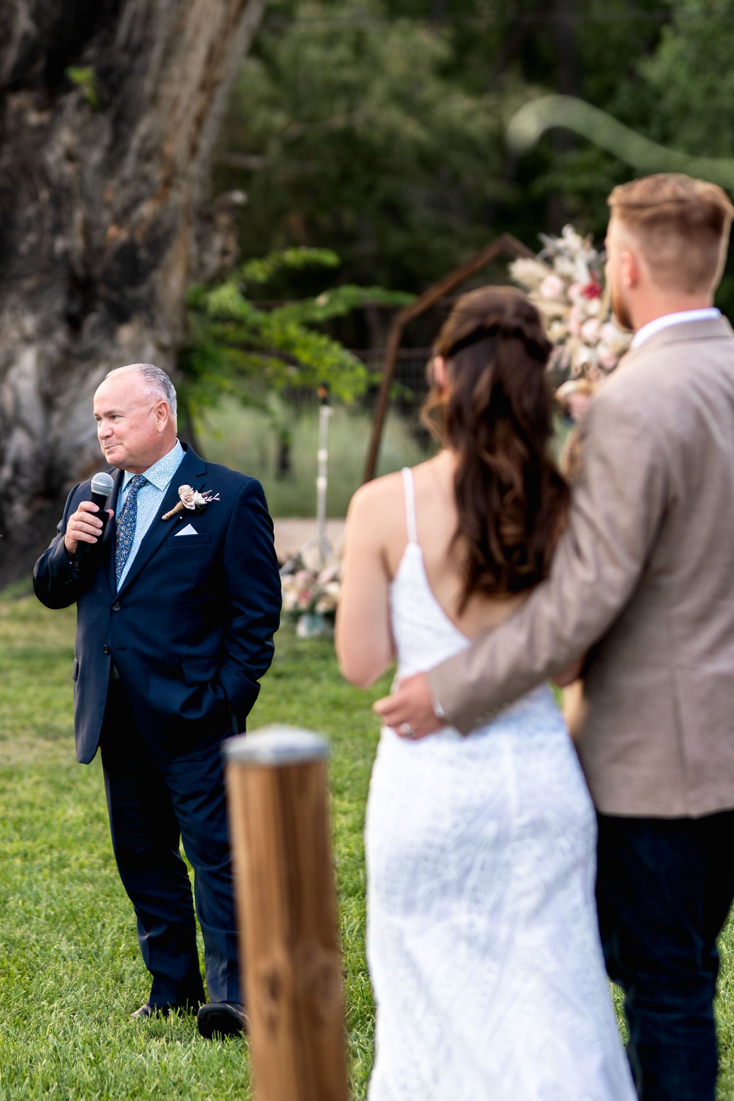 An officiant speaks into a microphone during an outdoor wedding ceremony with the couple standing nearby.