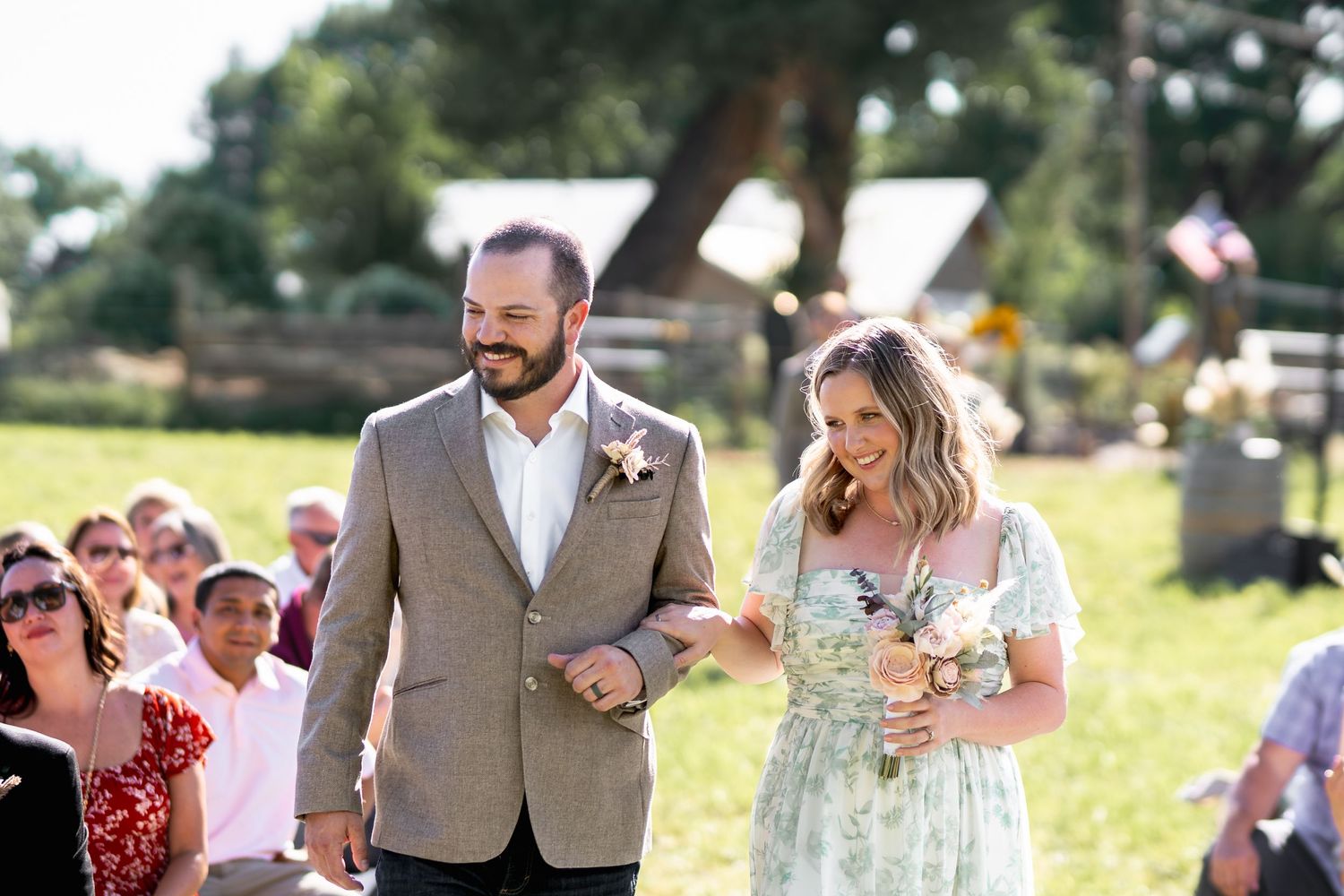 A couple walks down the aisle at an outdoor wedding ceremony while smiling guests look on.