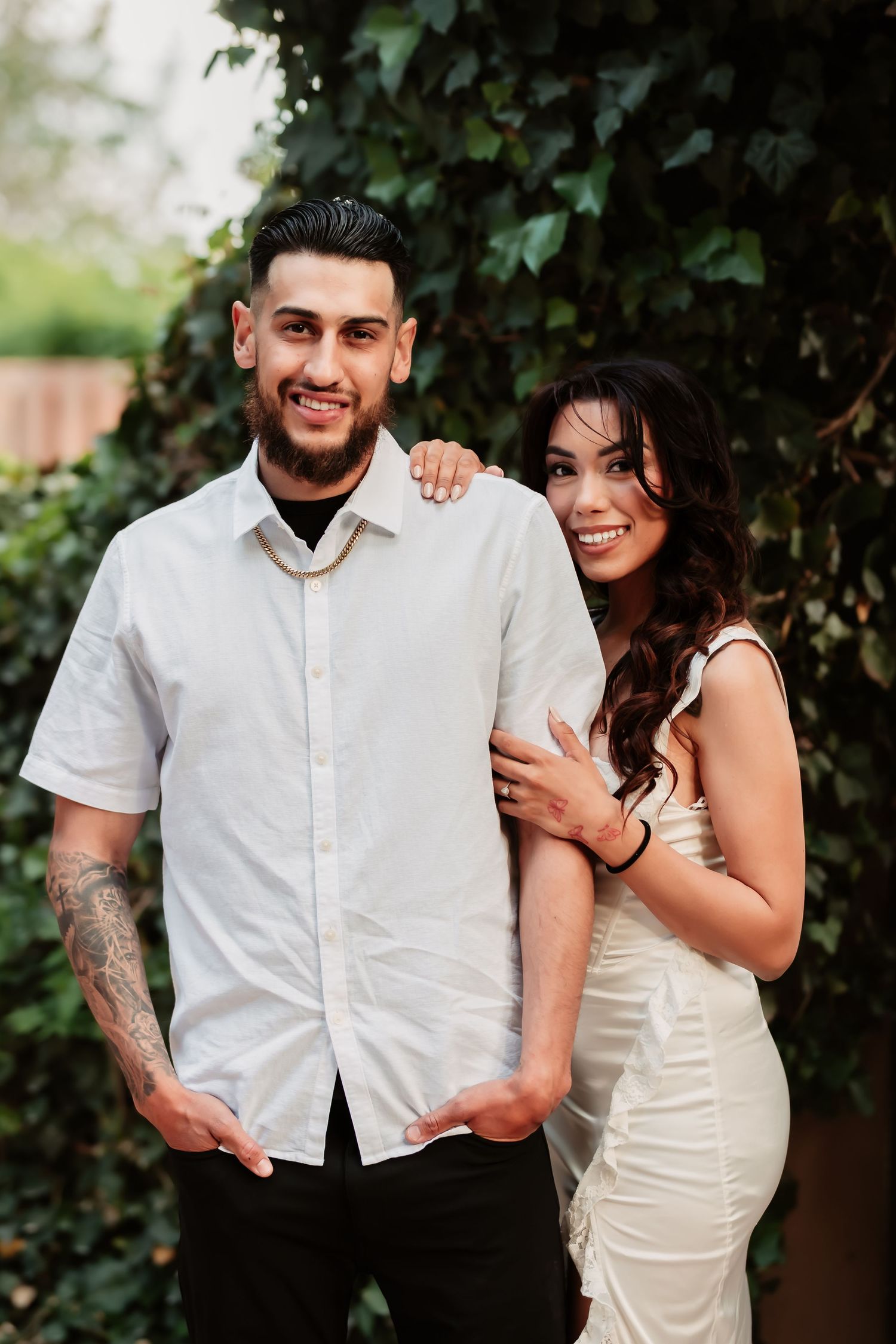 A couple in coordinating white outfits poses together against a leafy green backdrop for an engagement photo.