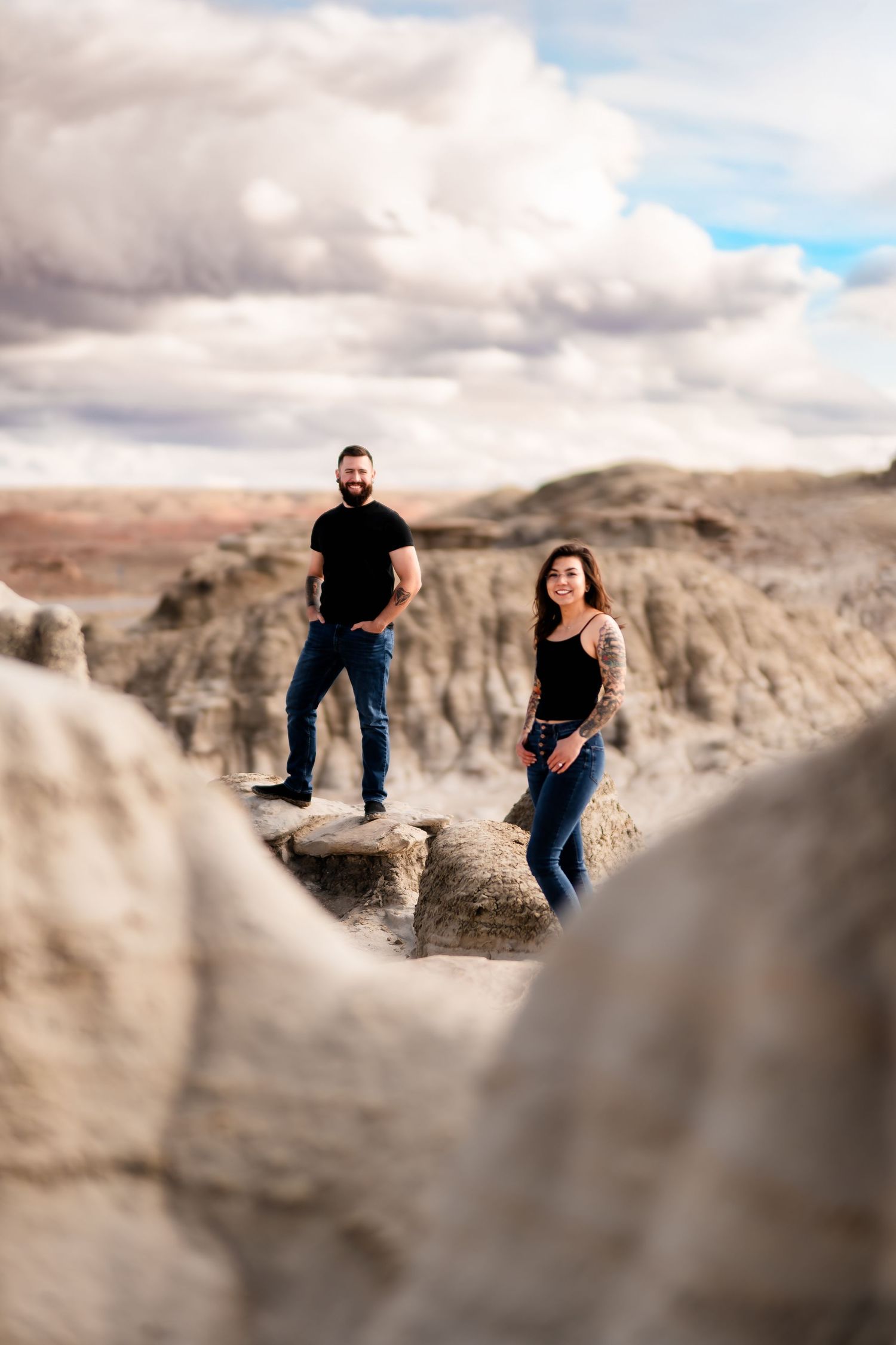 Two people stand on dramatic rock formations in a desert landscape under cloudy skies.