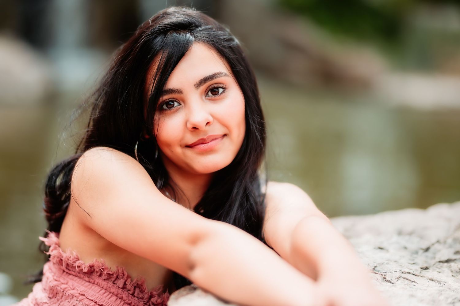 Portrait of a young woman with long dark hair smiling while resting on a stone ledge in an outdoor setting.