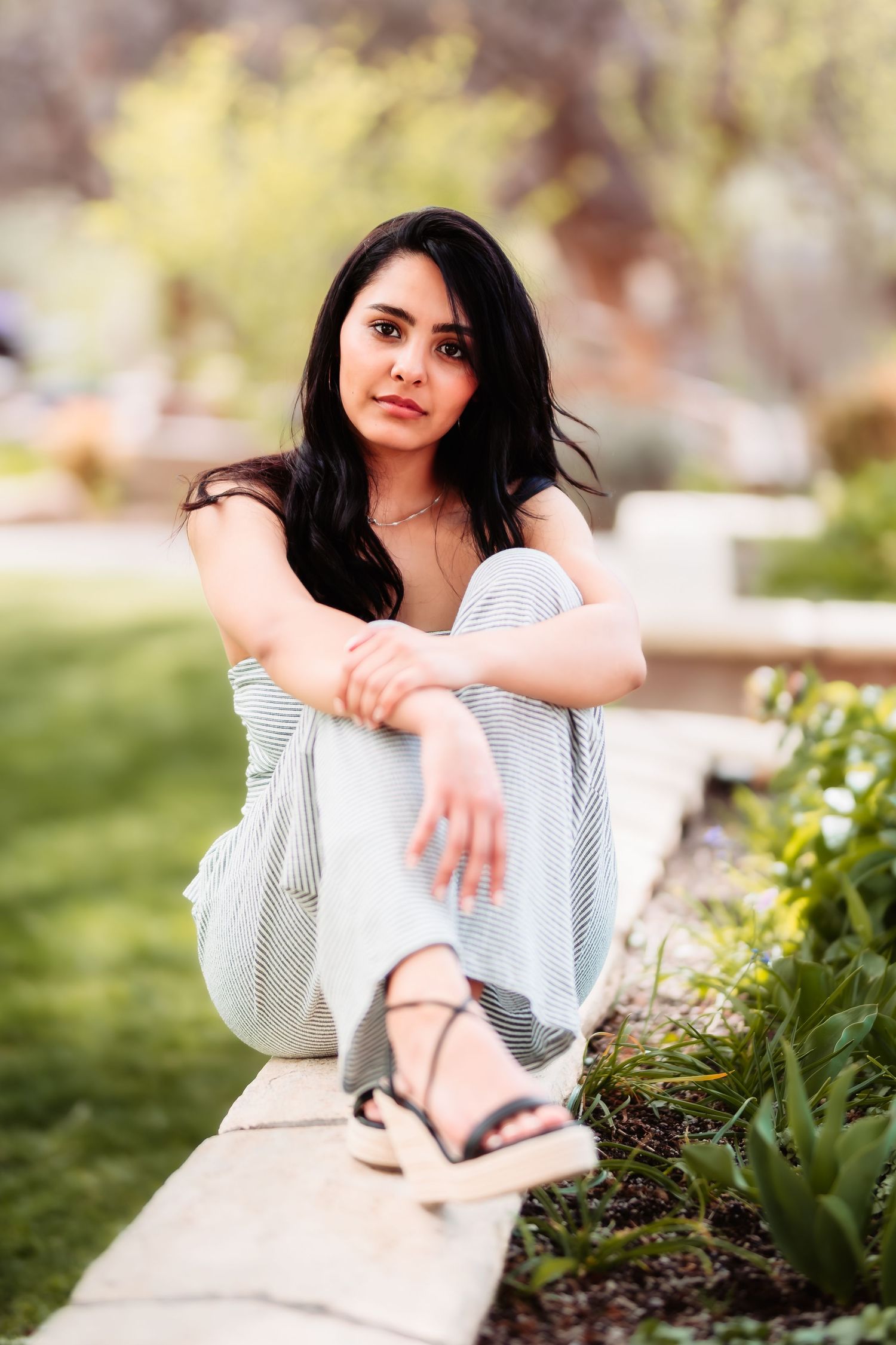 A woman in a casual striped outfit sits gracefully on a concrete ledge in an outdoor garden setting.