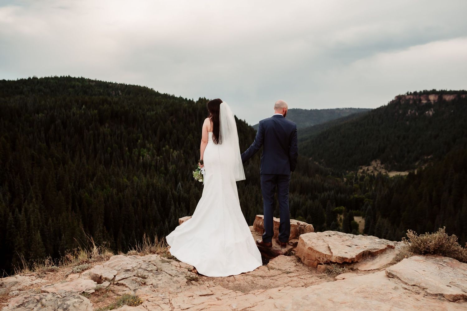 Newlywed couple stands on mountain cliff edge overlooking forested valley.