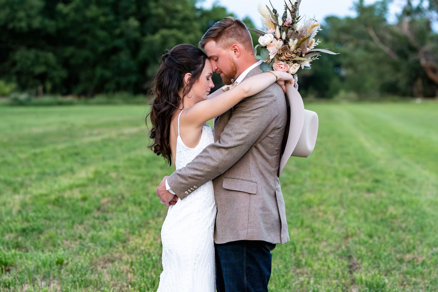 Newlyweds embrace in rustic outdoor setting with dried floral bouquet.