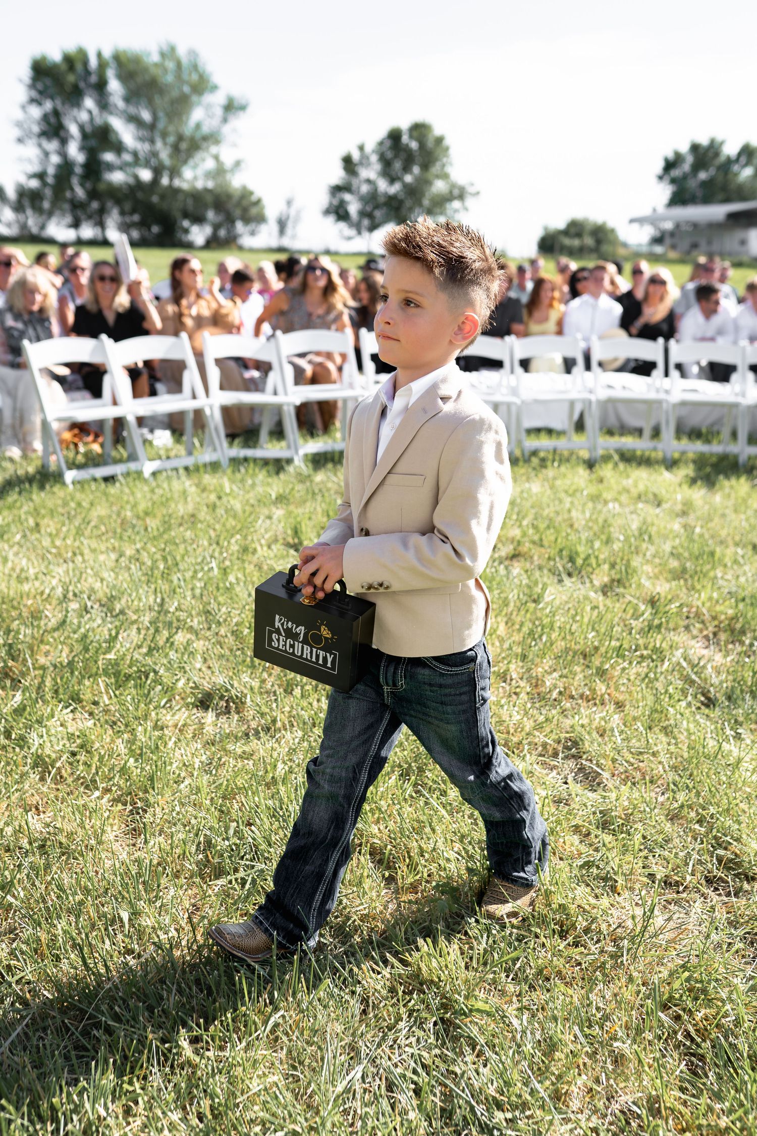 Young ring bearer in tan blazer and jeans carries a sign at outdoor wedding ceremony with white chairs visible.