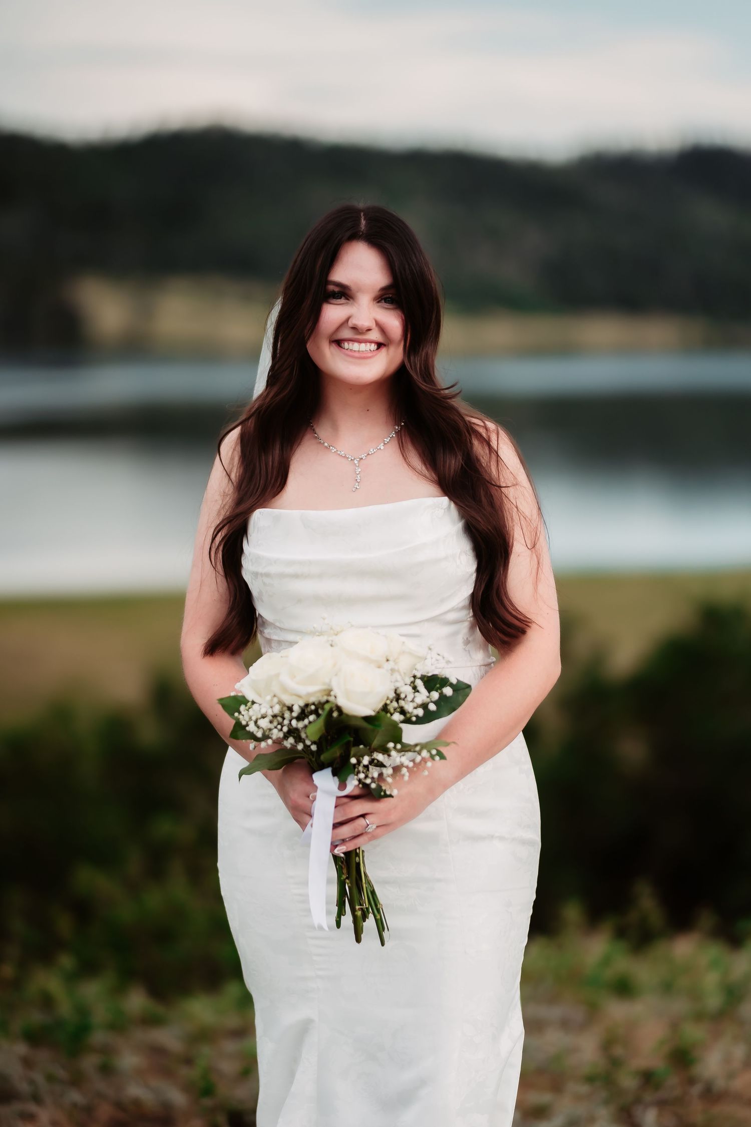 Bride smiling with white rose bouquet against scenic lake view.