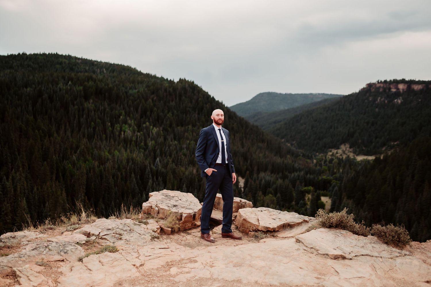 Groom in navy suit stands alone on rocky mountain overlook with forested valley below.