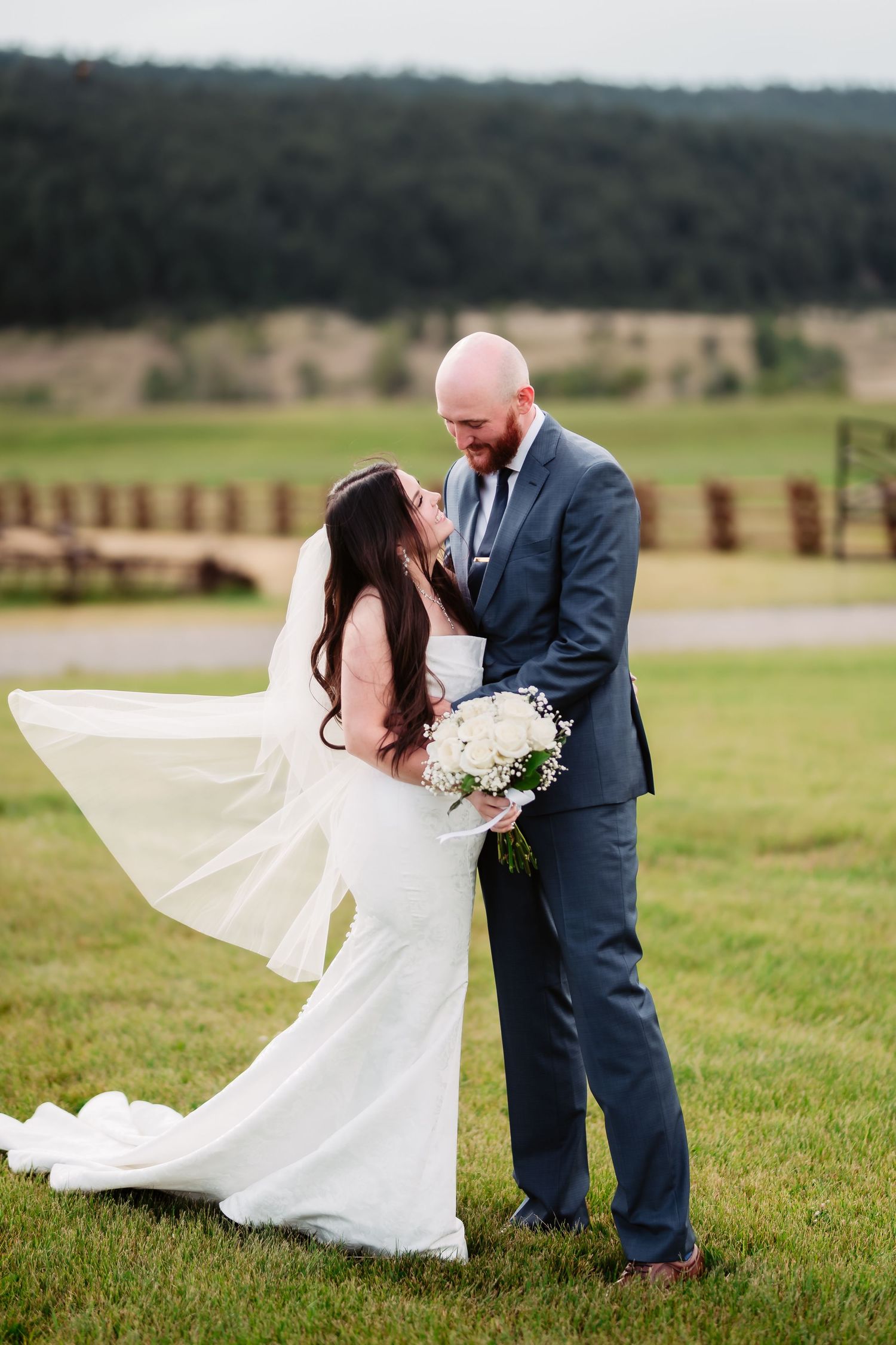Wedding couple poses in field as bride's white dress flows dramatically in the wind.