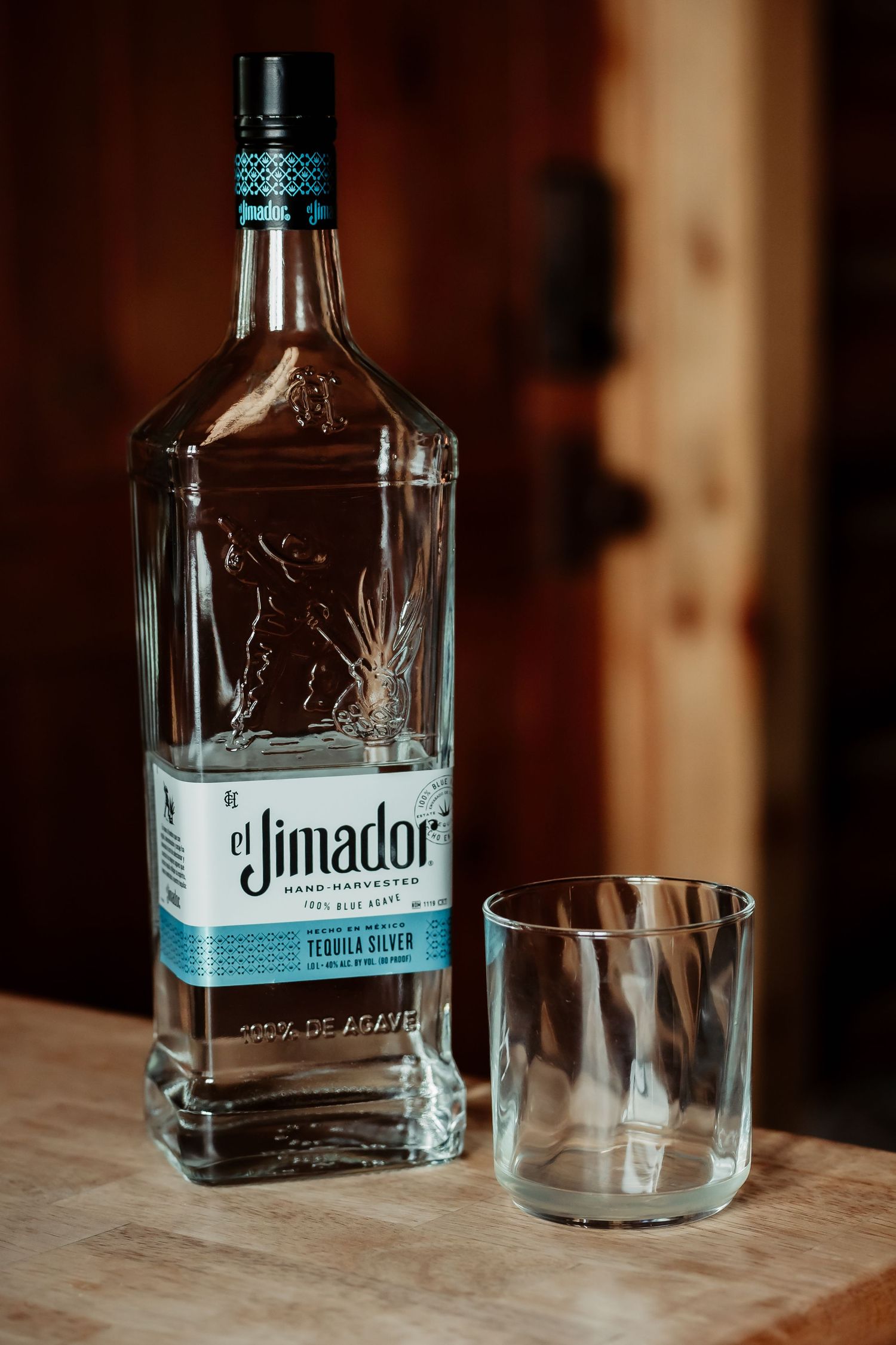 A bottle of Jimador tequila and an empty glass tumbler sit on a wooden surface in moody lighting.