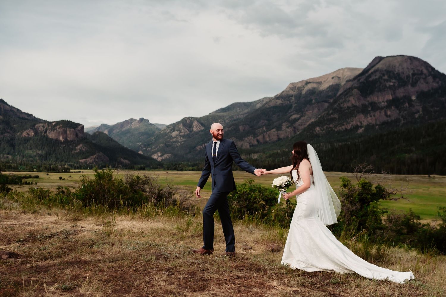 A couple shares a moment on a scenic mountain meadow with dramatic peaks in the background.