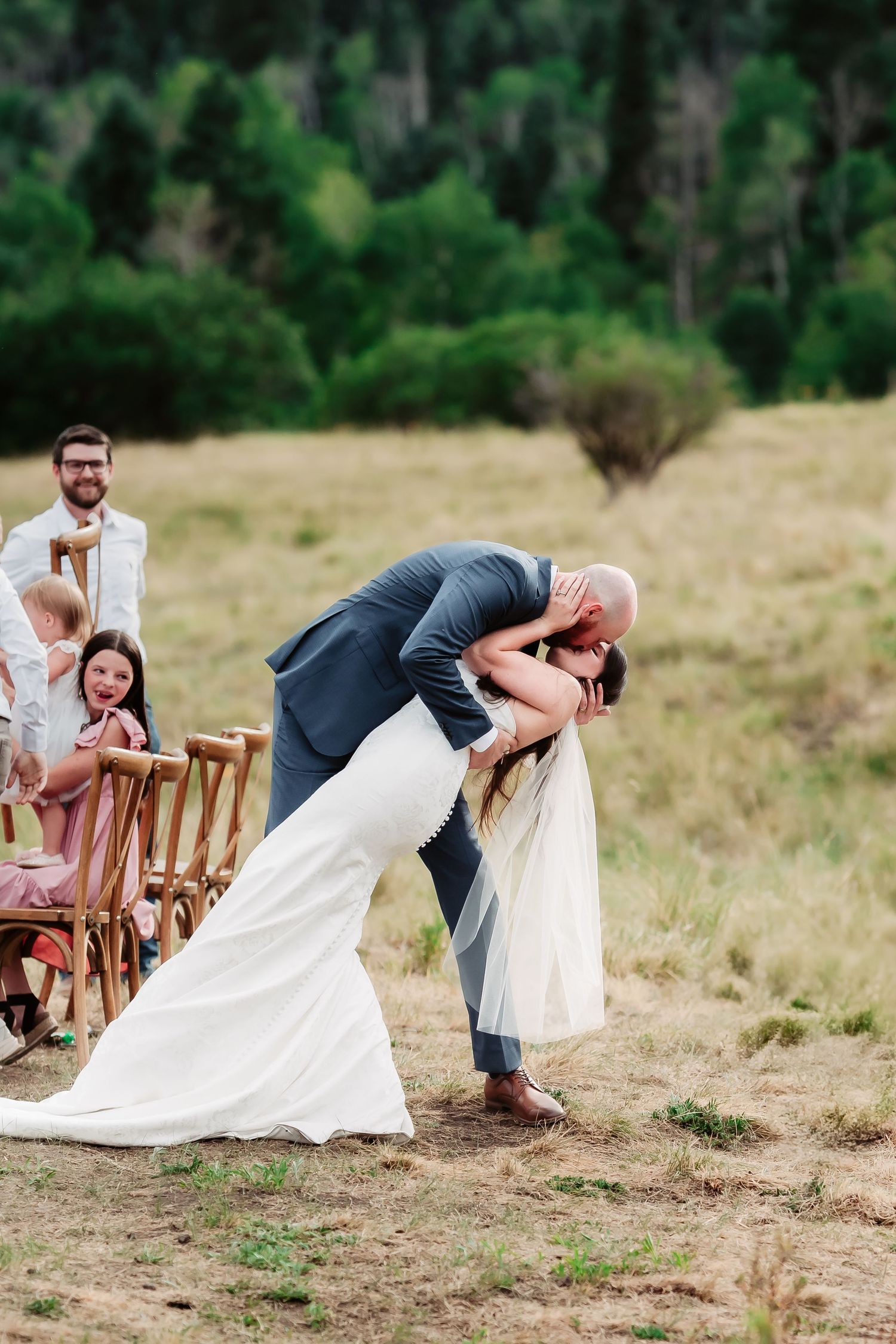 A dramatic wedding kiss dip pose during an outdoor ceremony with guests seated on wooden chairs.